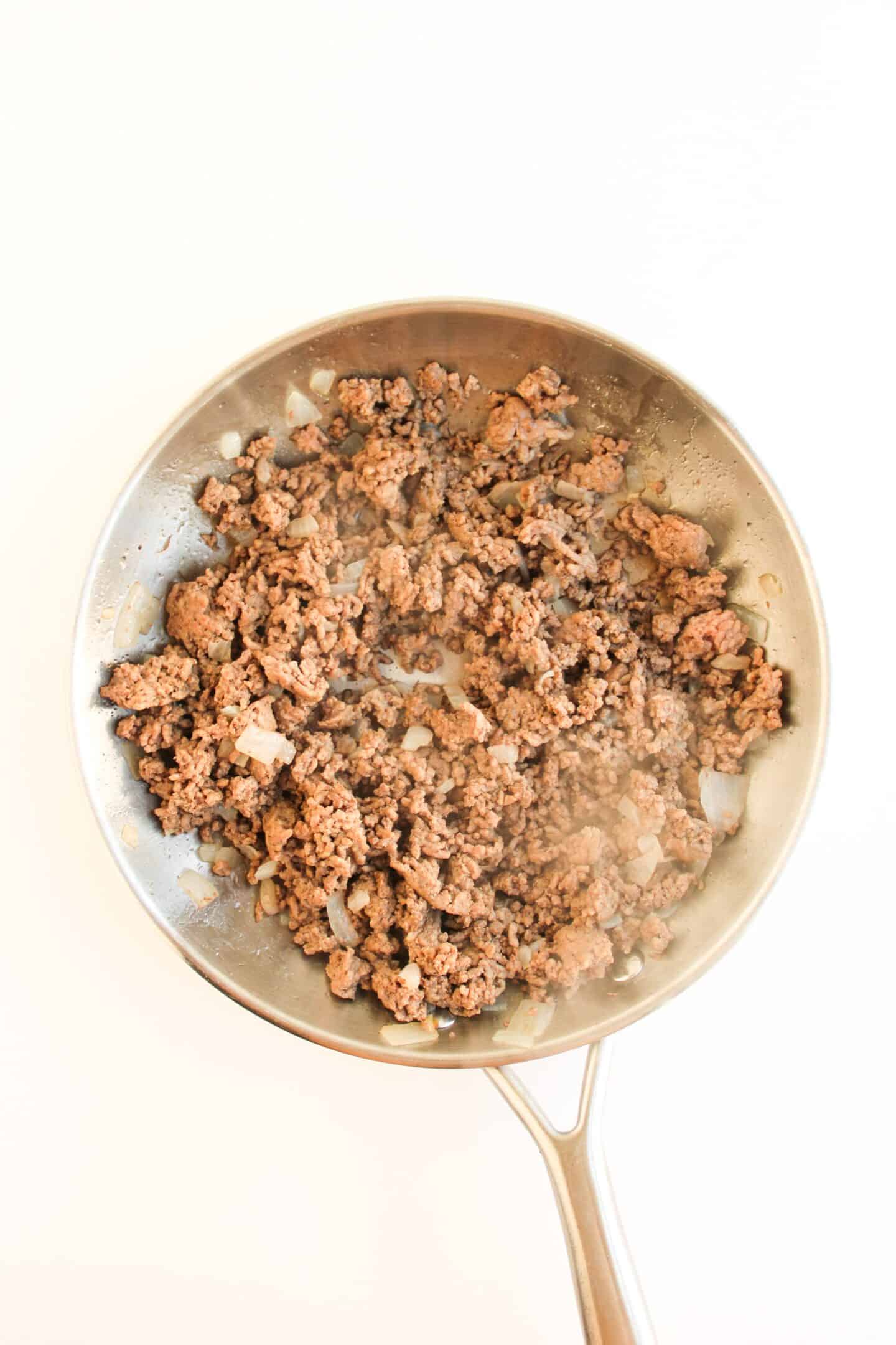 Cooked ground beef and chopped onions in a stainless steel skillet, perfect for your next Sloppy Joe recipe, on a white background.
