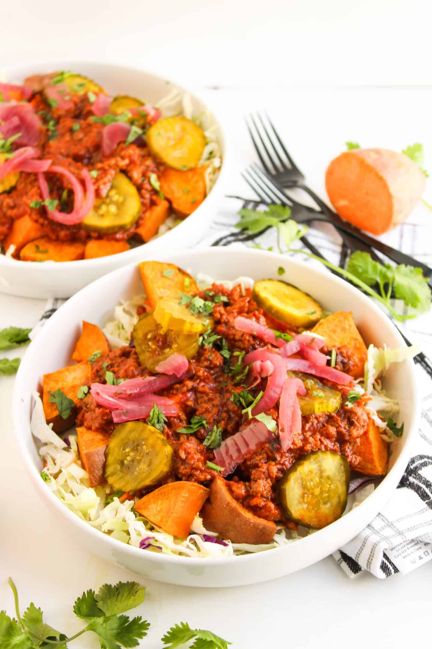 Two bowls filled with roasted sweet potatoes, ground meat in Sloppy Joe sauce, sliced pickles, pickled onions, and chopped greens on a bed of shredded cabbage, with utensils and cilantro nearby—Sloppy Joe Bowls reimagined.