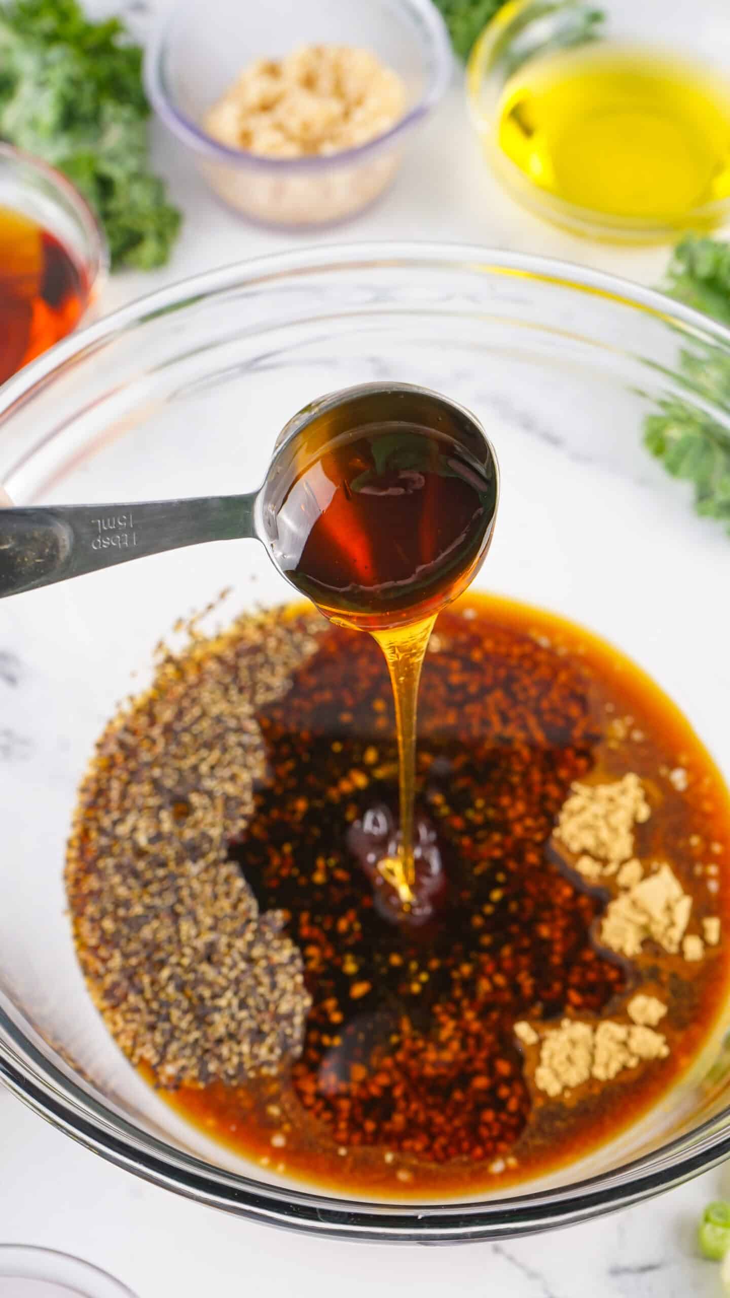 A metal tablespoon pours honey into a glass bowl of soy sauce, sesame seeds, chili flakes, and minced ginger—perfect for marinating Asian Style Pork Chops—with other fresh ingredients in small bowls nearby.
