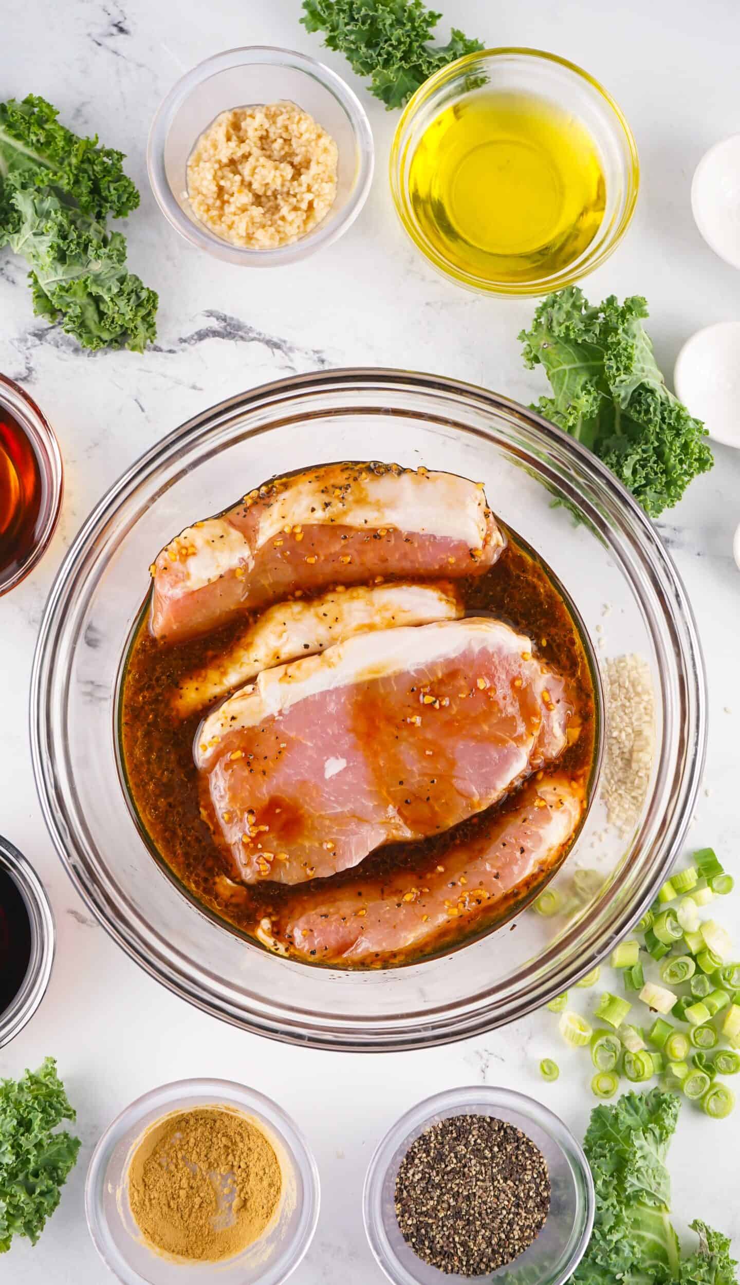 Three raw Asian Style Pork Chops marinating in a seasoned liquid in a glass bowl, surrounded by chopped green onions, kale, spices, oil, and other ingredients on a white surface.