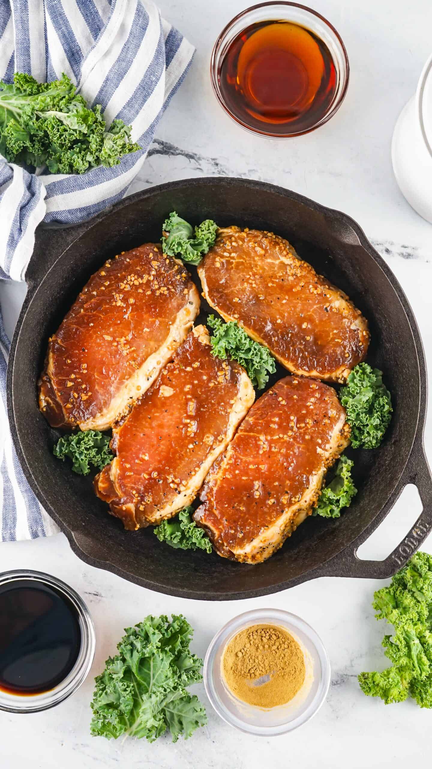 Four seasoned Asian Style Pork Chops are arranged in a cast iron skillet with fresh kale, surrounded by small bowls of ingredients and a striped towel on a white surface.