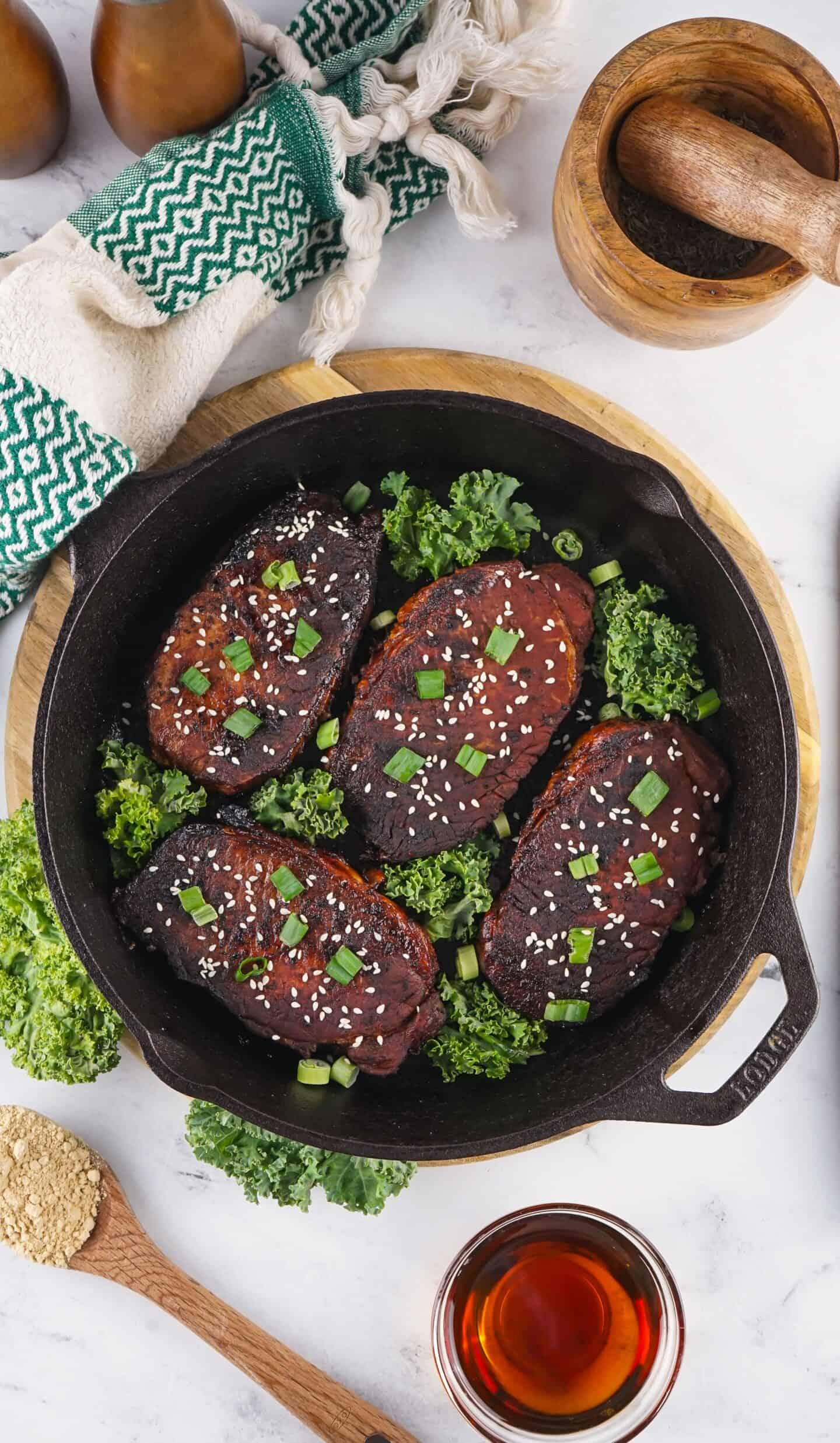 Four seared, glazed tempeh steaks topped with sesame seeds and chopped green onions in a cast iron skillet, surrounded by curly kale—an Asian Style Pork Chops-inspired dish—on a white countertop with kitchen utensils.