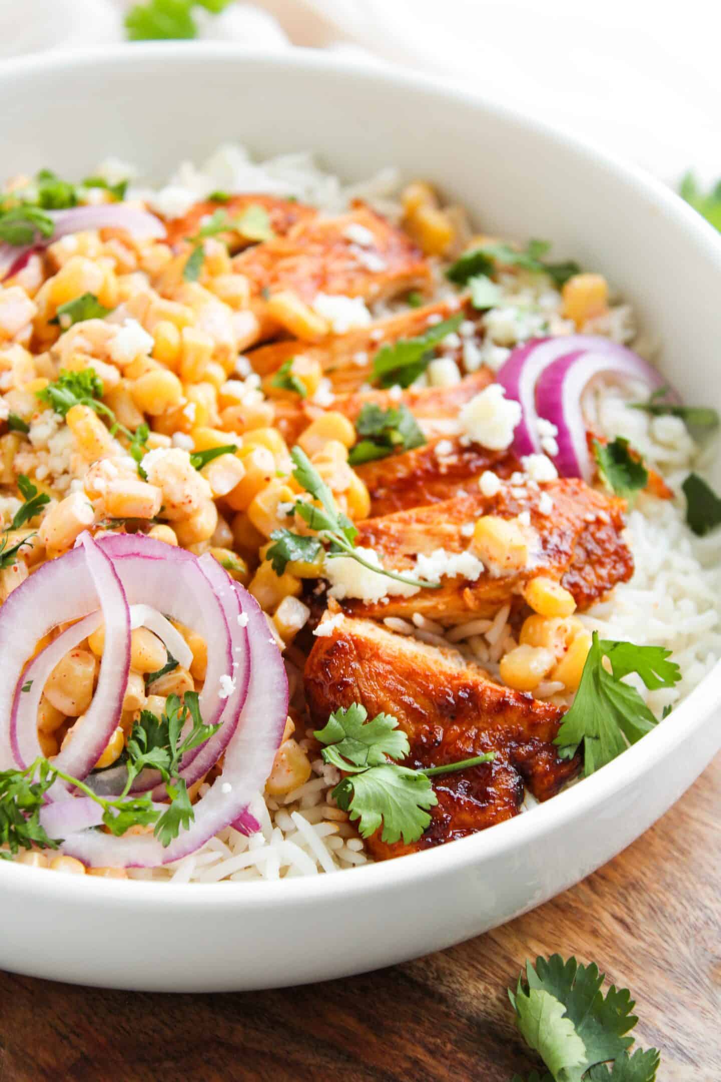 A bowl of white rice topped with grilled chicken, corn, sliced red onions, cilantro, and crumbled cheese—our Chicken Street Corn Bowls bring bold, fresh flavors to every bite.