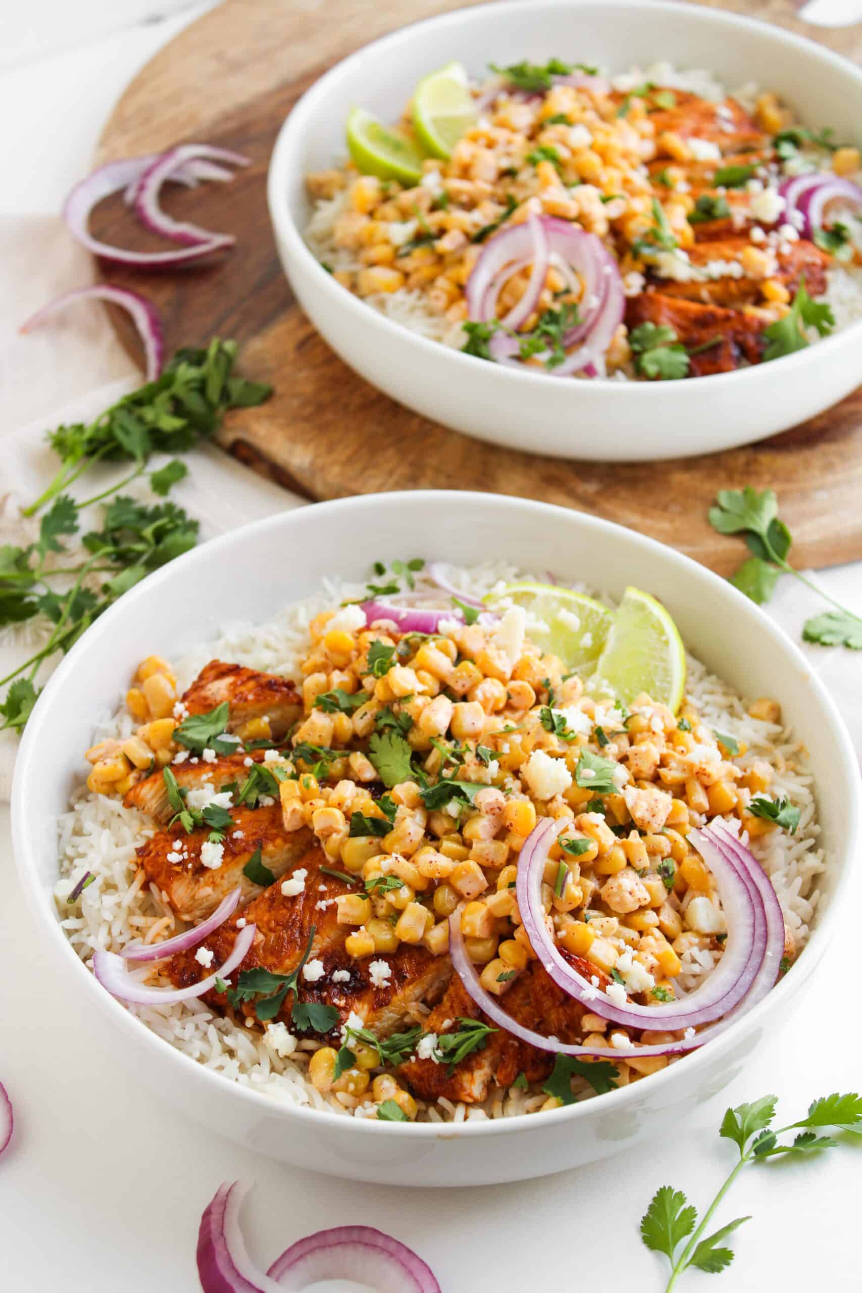 Two white bowls filled with rice, juicy grilled chicken, street corn, red onion slices, chopped herbs, lime wedges, and crumbled cheese are set on a table with cilantro and onion garnish.
