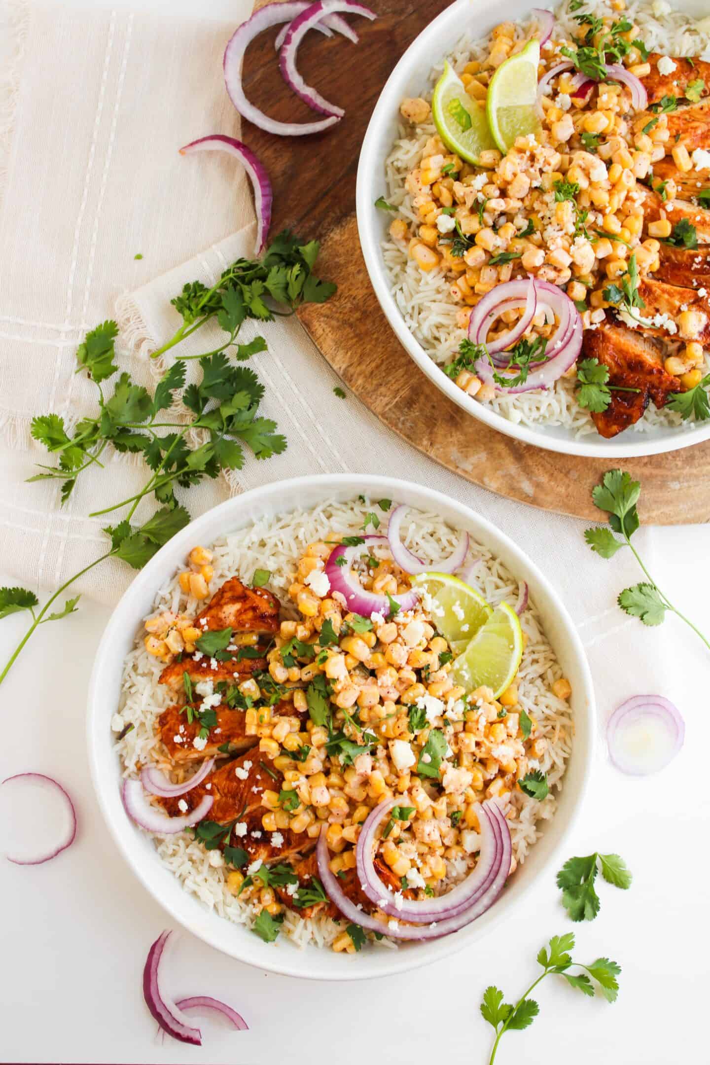 Two white bowls filled with rice, grilled chicken, street corn, red onion slices, lime wedges, cilantro, and cheese crumbles sit on a white surface with scattered cilantro and onions.