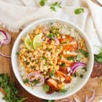A bowl of Chicken Street Corn Bowls topped with grilled chicken, corn salsa, red onion slices, lime wedges, crumbled cheese, and fresh cilantro, placed on a wooden board with herbs and a napkin.