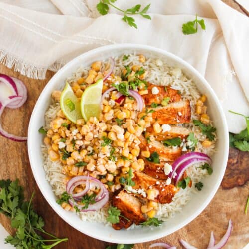 A bowl of Chicken Street Corn Bowls topped with grilled chicken, corn salsa, red onion slices, lime wedges, crumbled cheese, and fresh cilantro, placed on a wooden board with herbs and a napkin.