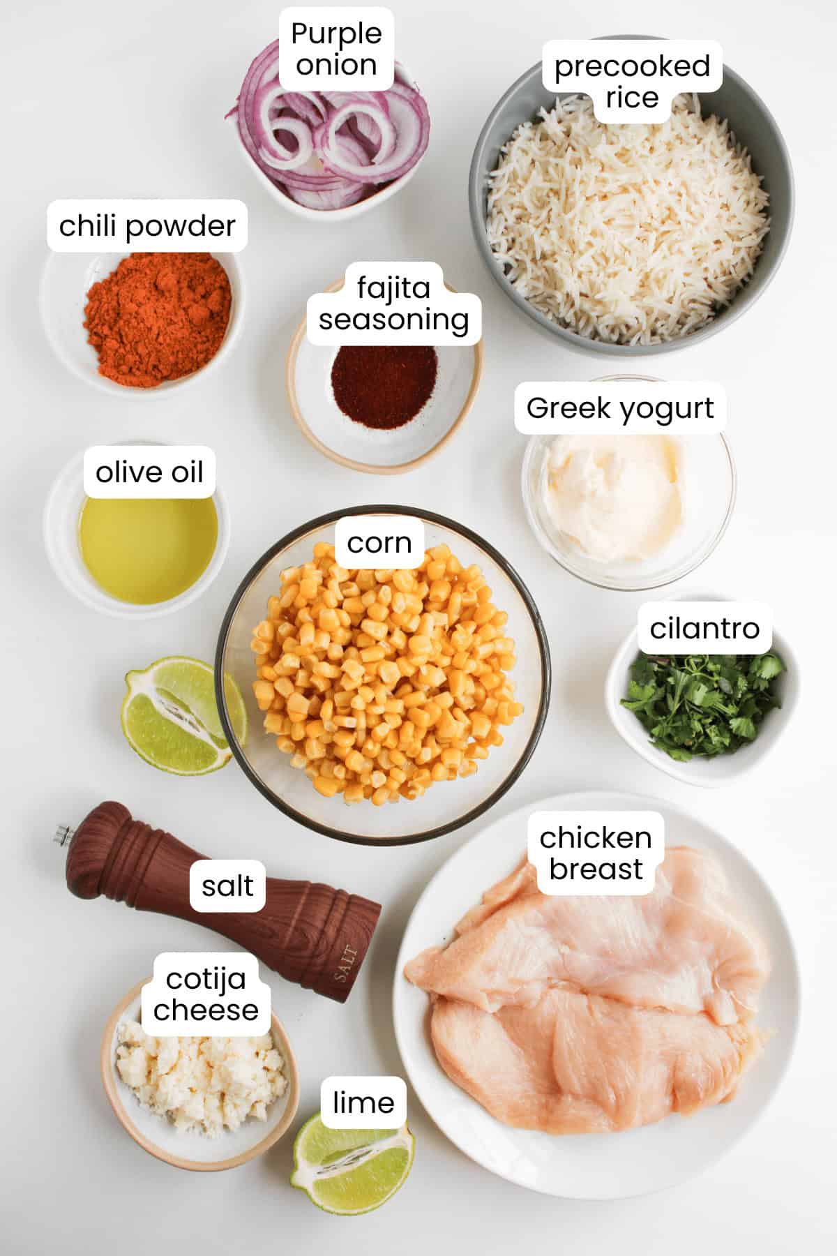 Ingredients for delicious street corn chicken bowls are arranged on a white surface, including chicken breast, corn, precooked rice, Greek yogurt, spices, purple onion, cilantro, cotija cheese, lime, olive oil, and salt.