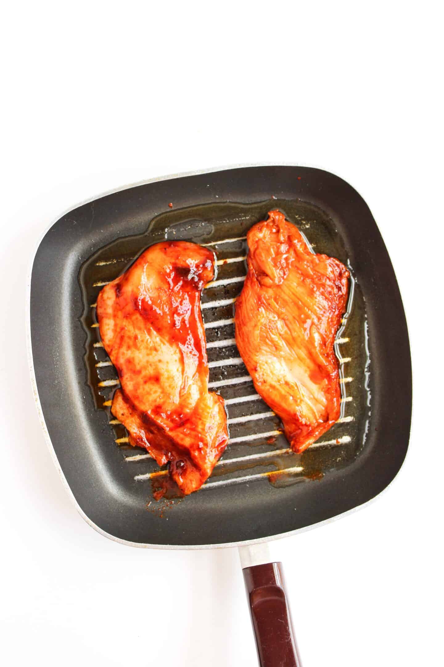 Two pieces of marinated chicken breast are cooking on a black grill pan against a white background, perfect for adding to flavorful Chicken Street Corn Bowls.