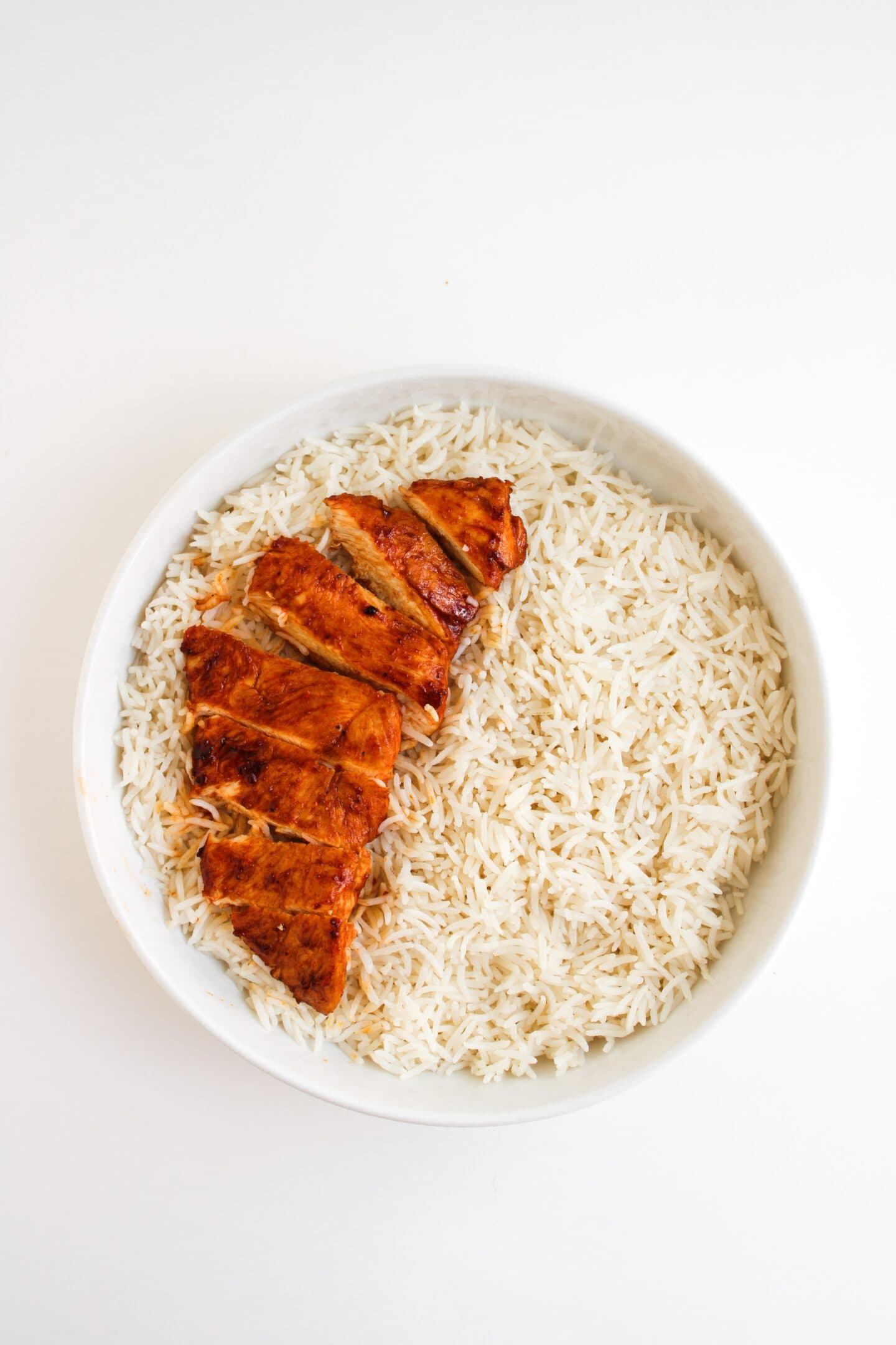 A white bowl filled with plain white rice and tender sliced grilled chicken breast arranged on top, set against a white background.
