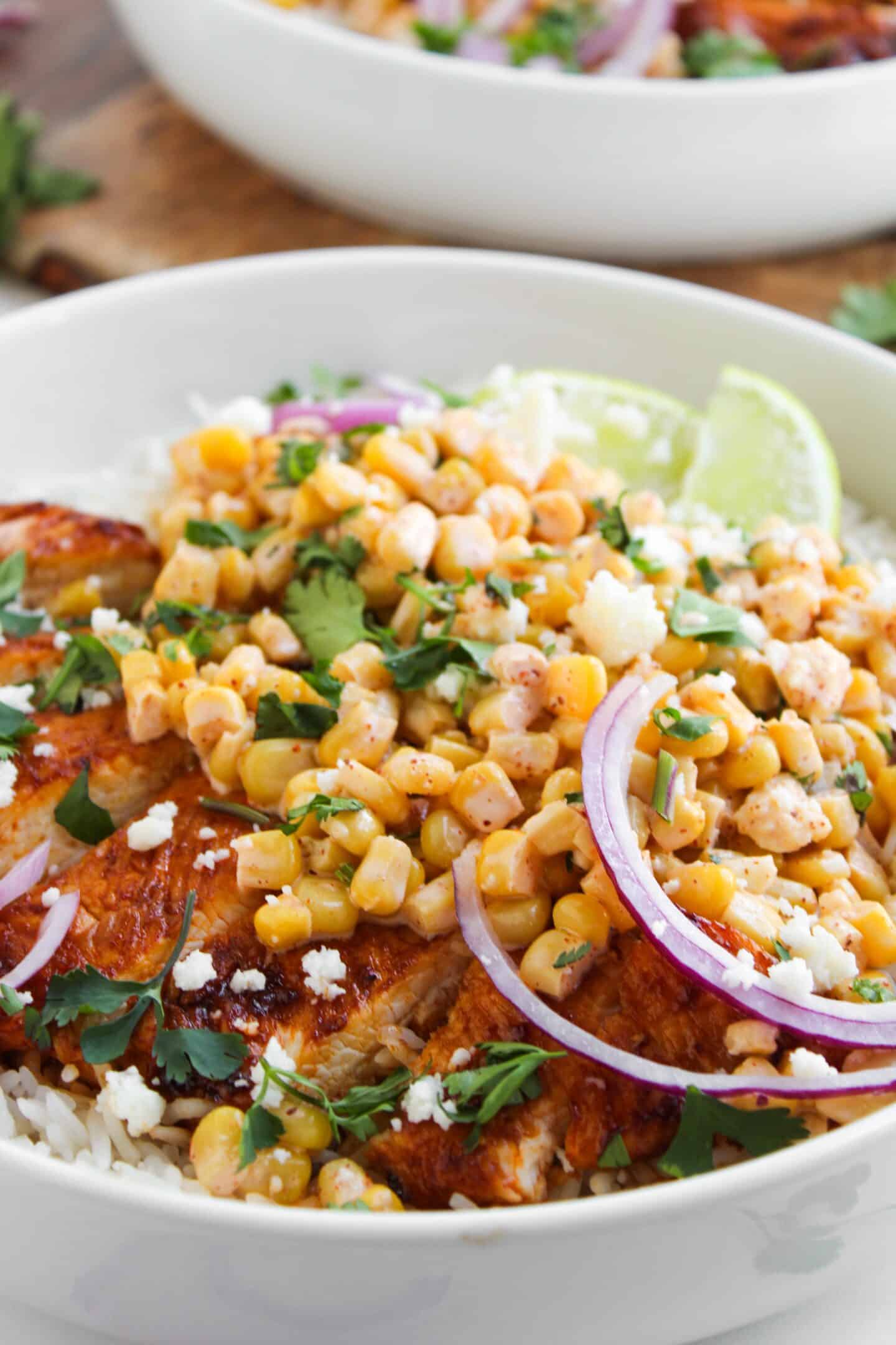 A bowl of rice topped with grilled chicken, corn, red onion slices, cilantro, crumbled cheese, and lime wedges—your perfect Chicken Street Corn Bowl for a fresh and flavorful meal.