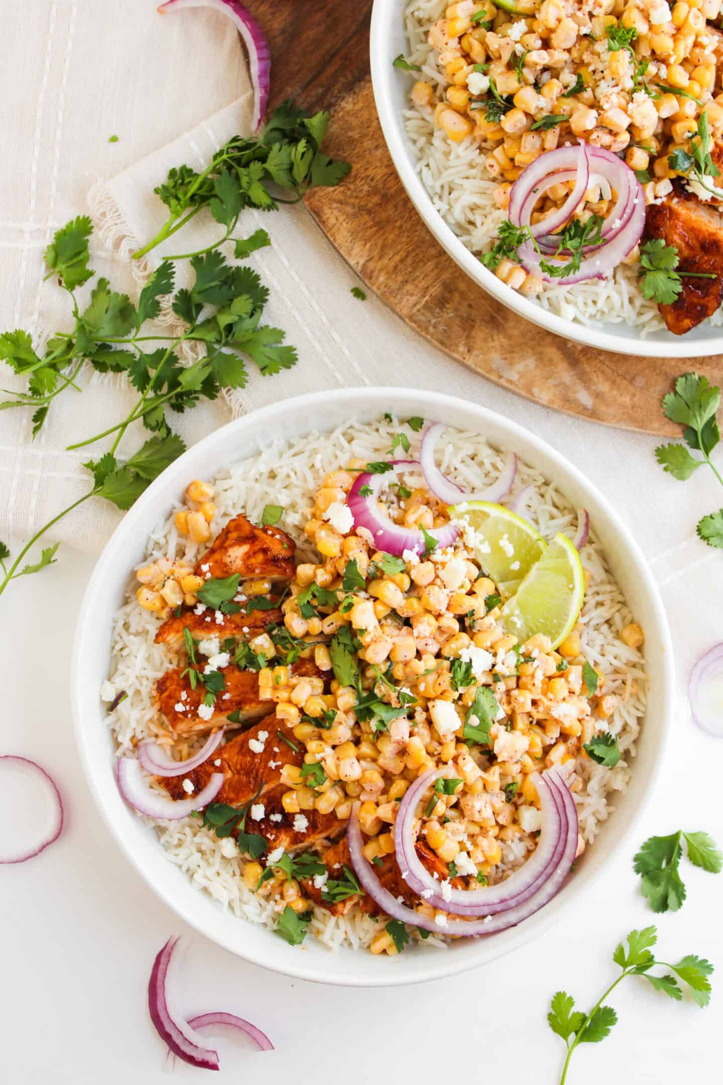 A Chicken Street Corn Bowl filled with rice, grilled chicken, corn, red onion slices, cilantro, a lime wedge, and crumbled cheese. Another Chicken Street Corn Bowl sits partially visible beside it.