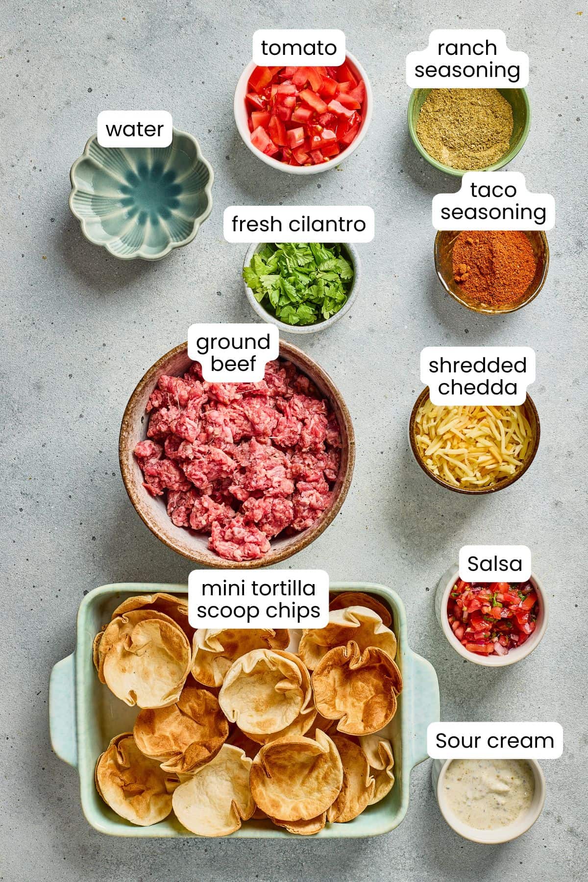 Ingredients for taco cups are arranged on a gray surface—ground beef, mini tortilla scoop chips, tomato, fresh cilantro, shredded cheddar, salsa, seasonings, water, and sour cream—making it easy to follow any recipe template.