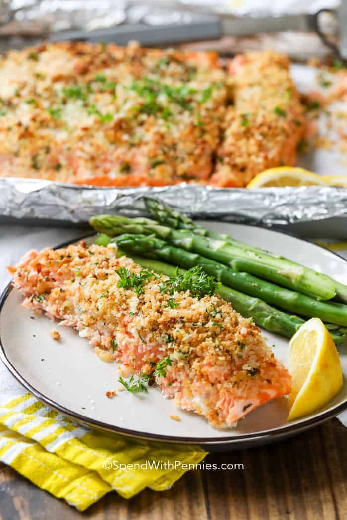 A plate with a serving of baked salmon topped with breadcrumbs and herbs, steamed asparagus, and a lemon wedge—perfect for cozy Fall Salmon Recipes. More salmon on foil is visible in the background.