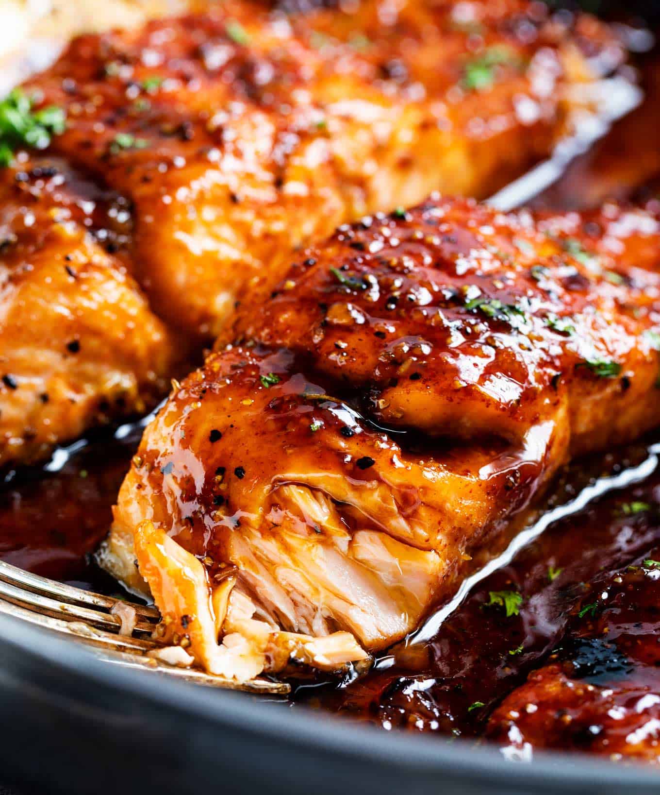 Close-up of glazed, cooked salmon fillets in a pan, perfect for fall salmon recipes, topped with herbs and coated in a shiny sauce, with a fork flaking one piece.