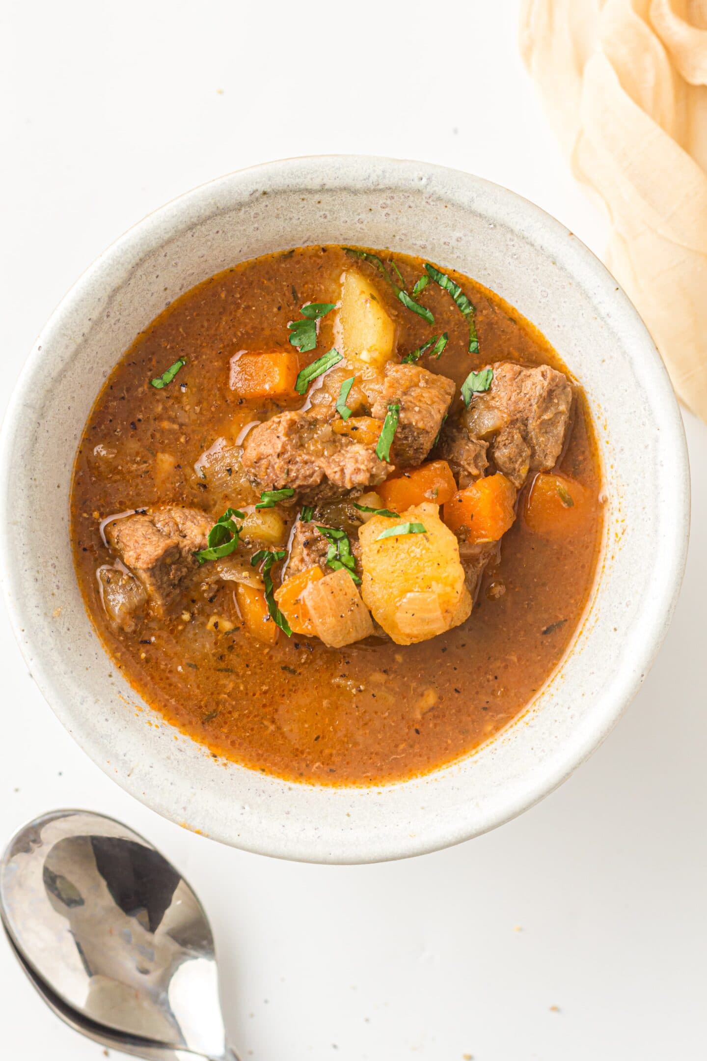 A bowl of hearty Beef Stew with chunks of beef, potatoes, carrots, and herbs in a rich brown broth sits next to a spoon on a white surface—perfectly cooked in the Instant Pot for ultimate convenience.