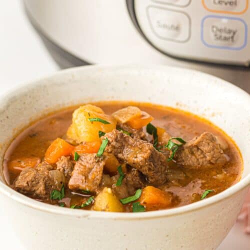 A bowl of hearty Beef Stew with carrots and potatoes, garnished with herbs, sits in front of an Instant Pot.