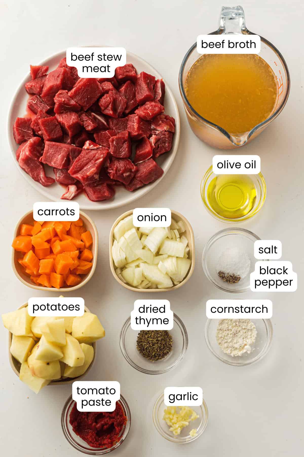 Overhead view of raw Beef Stew ingredients for an Instant Pot, including beef stew meat, beef broth, olive oil, carrots, onion, potatoes, tomato paste, dried thyme, garlic, cornstarch, salt, and black pepper.