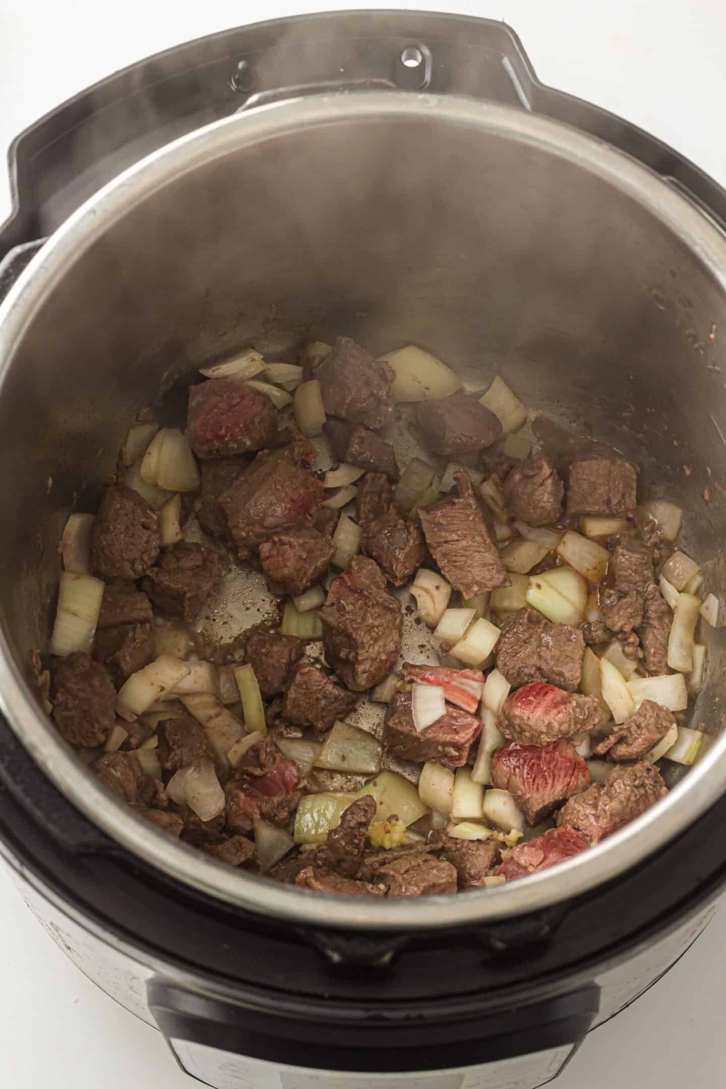 Chunks of beef and diced onions being sautéed inside an open Instant Pot, the perfect start for a hearty Beef Stew.