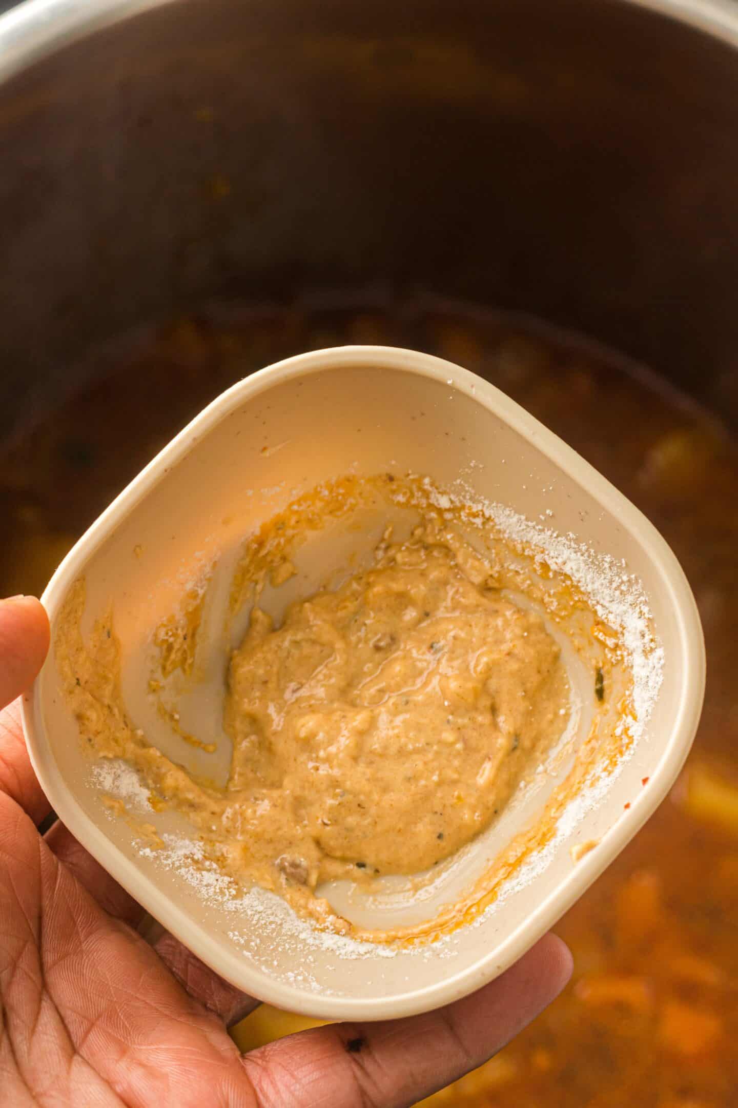A hand holding a small beige bowl with a mixture of spices and liquid, positioned above an Instant Pot full of savory beef stew.