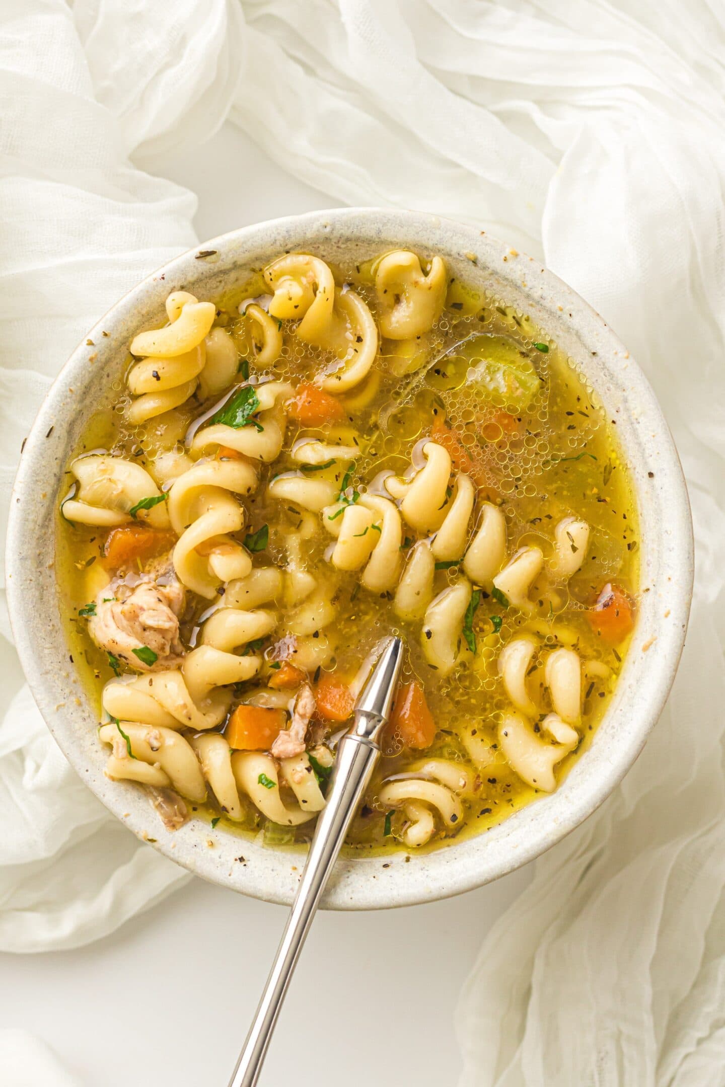 A bowl of rotini pasta soup with vegetables, herbs, and pieces of Instant Pot Chicken, served with a spoon on a white surface and a white cloth nearby.