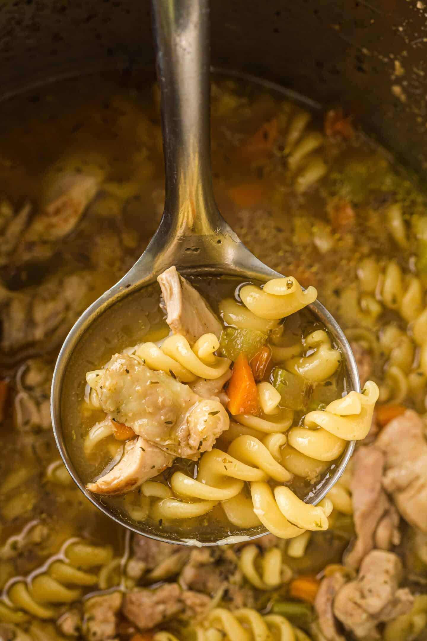 A metal ladle holds rotini pasta, chunks of chicken, and vegetables above a pot of Chicken Noodle Soup simmering in an Instant Pot.