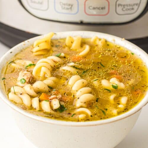 A bowl of rotini pasta soup with vegetables sits in front of an Instant Pot, bringing a modern twist to classic Chicken Noodle Soup.