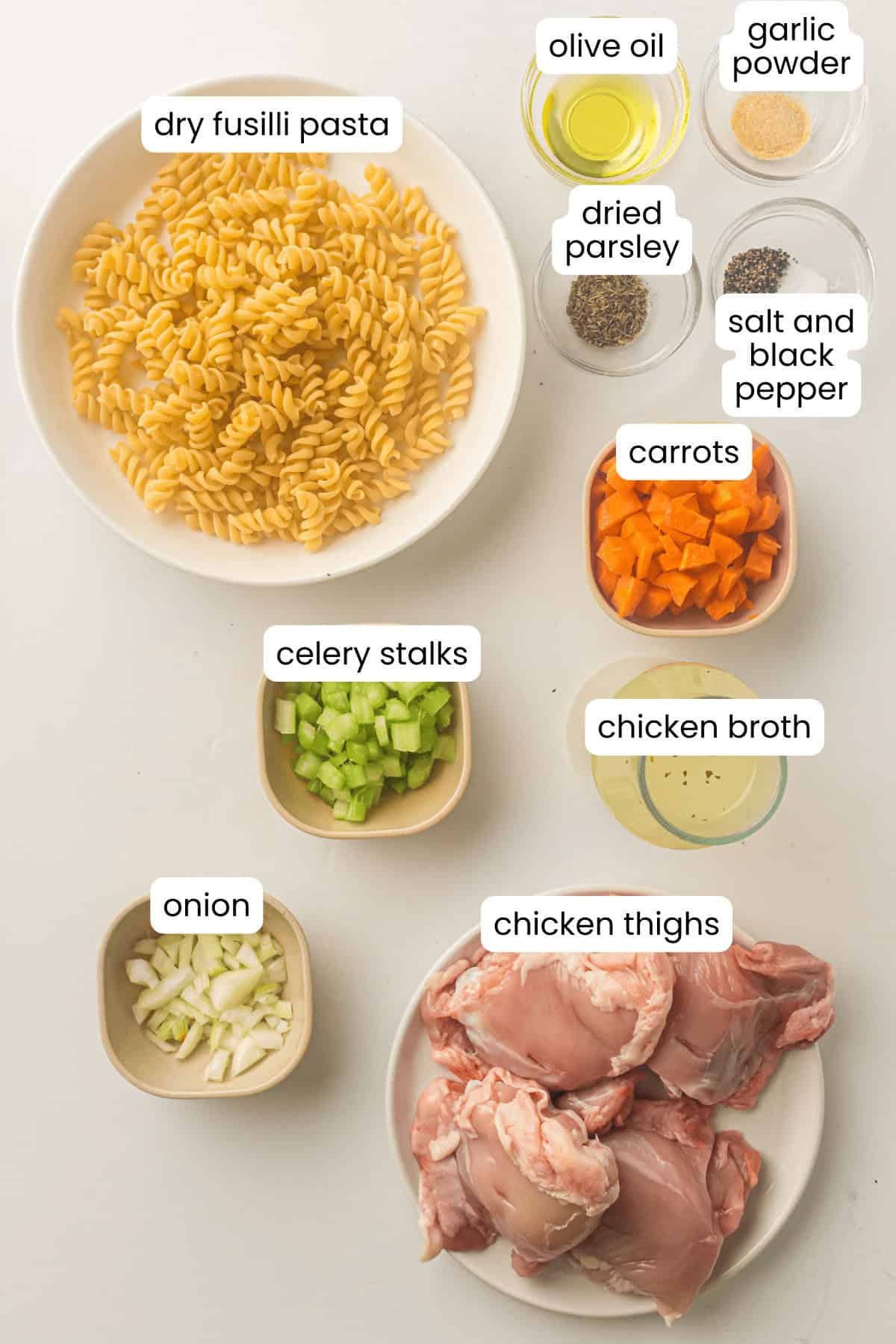Ingredients for an Instant Pot chicken noodle soup are laid out on a white surface, featuring fusilli, chicken thighs, broth, onion, celery, carrots, parsley, olive oil, garlic powder, salt, and pepper.