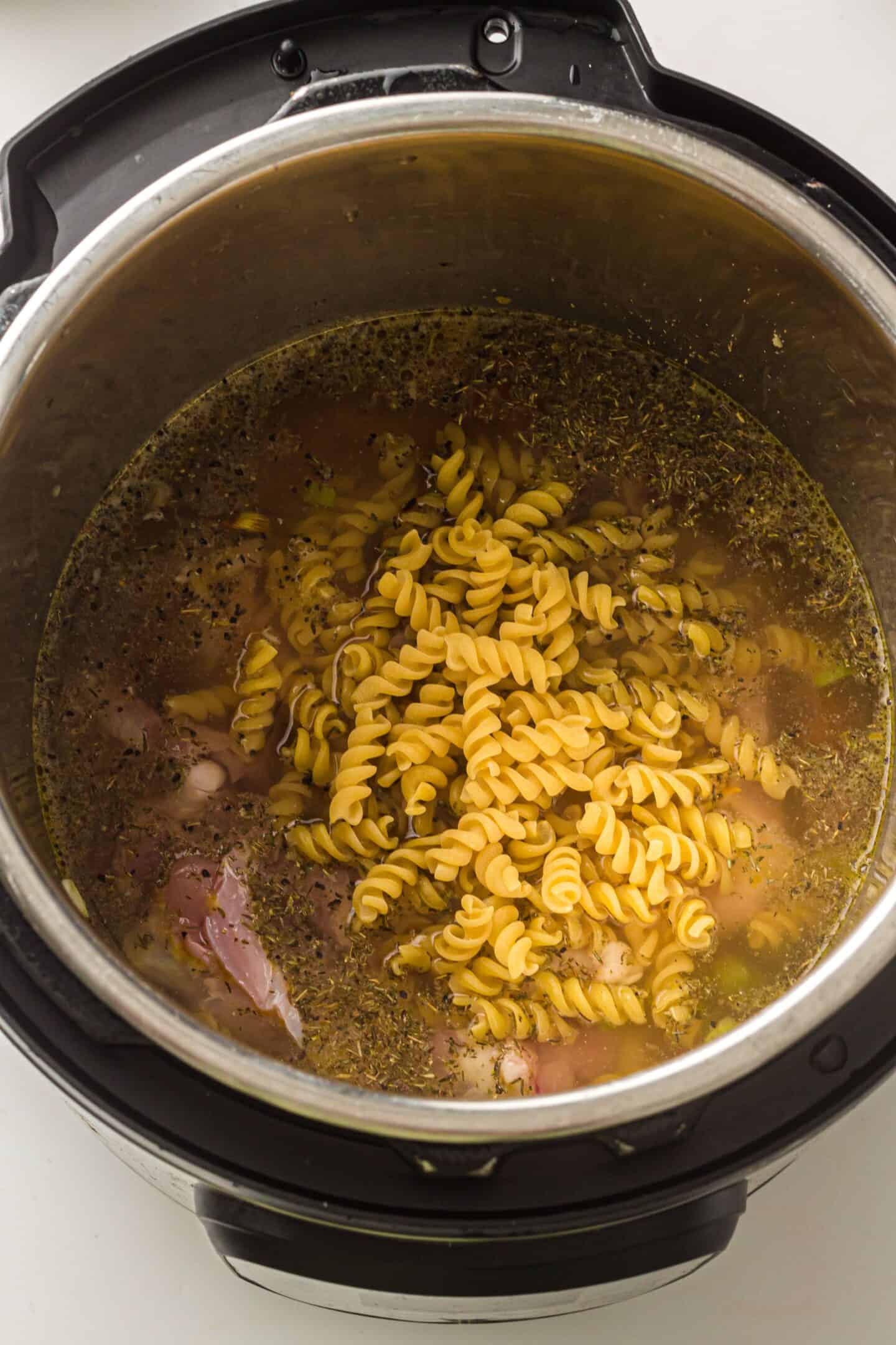 Uncooked rotini pasta, raw chicken, broth, and herbs in an Instant Pot await transformation into a comforting Chicken Noodle Soup.