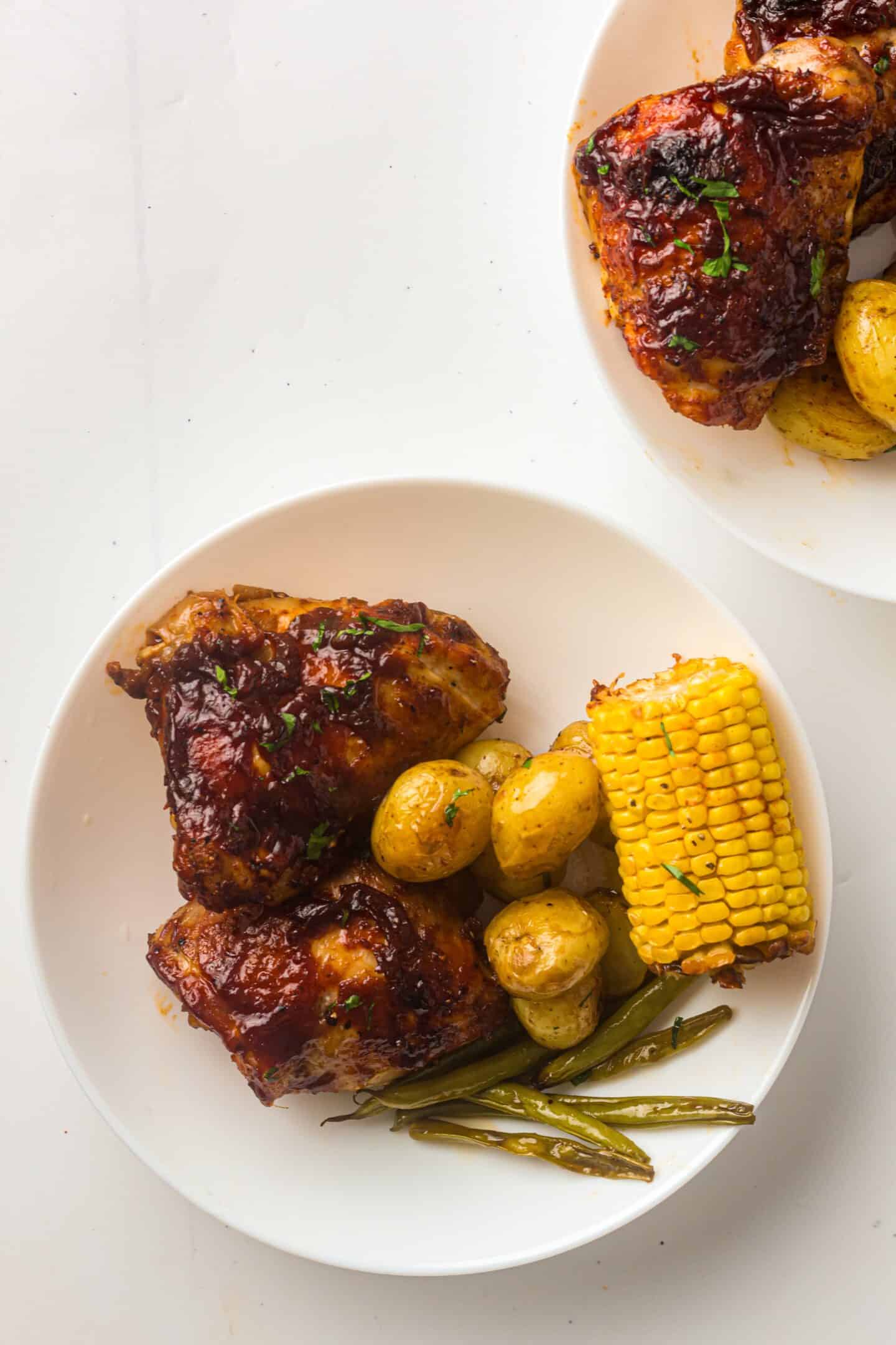 A white plate with BBQ chicken thighs, roasted baby potatoes, green beans, and a piece of corn on the cob.