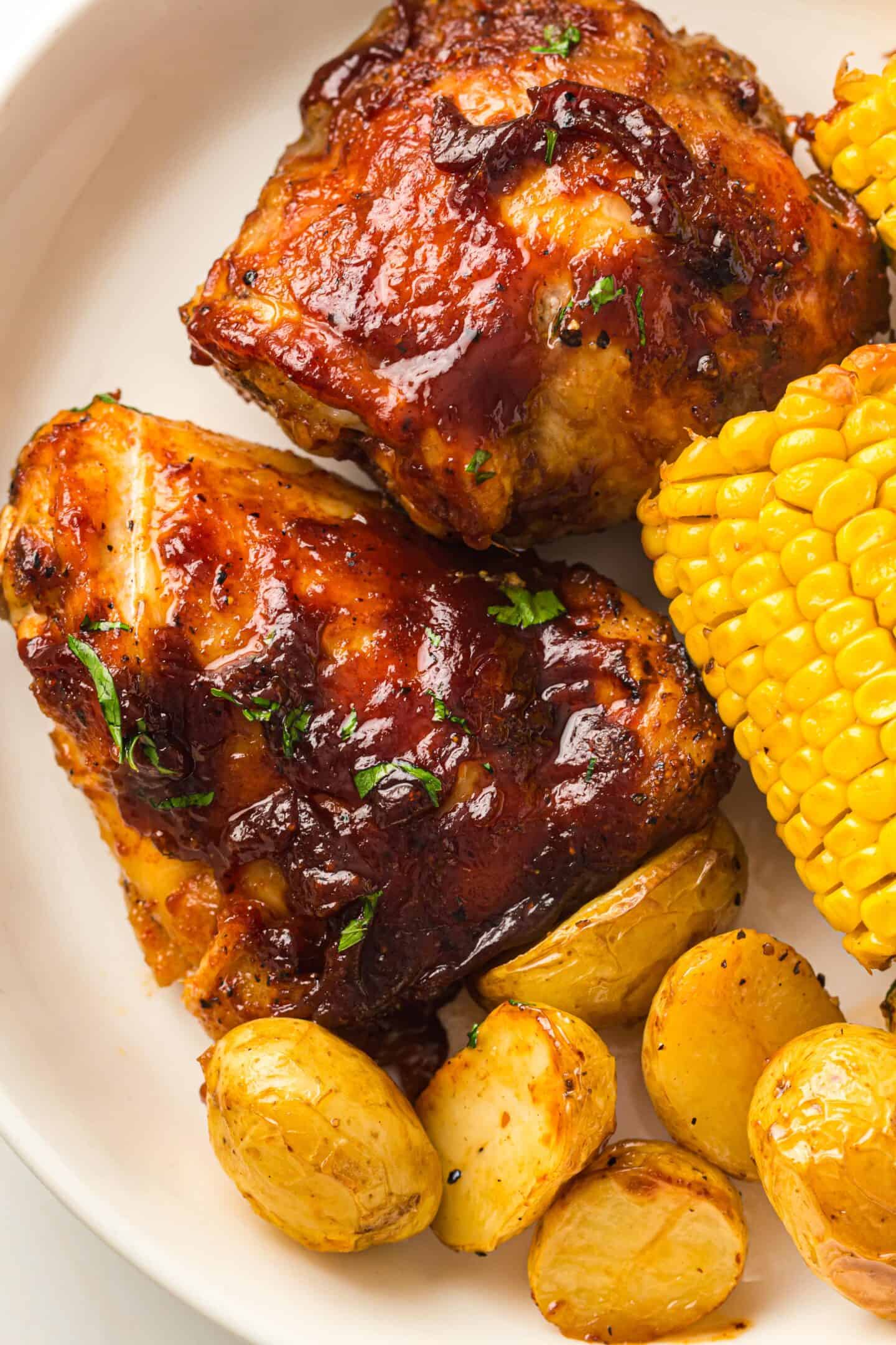 Plate with two pieces of juicy BBQ Chicken Thighs, roasted potatoes, and a section of corn on the cob—all perfectly cooked on a single sheet pan.
