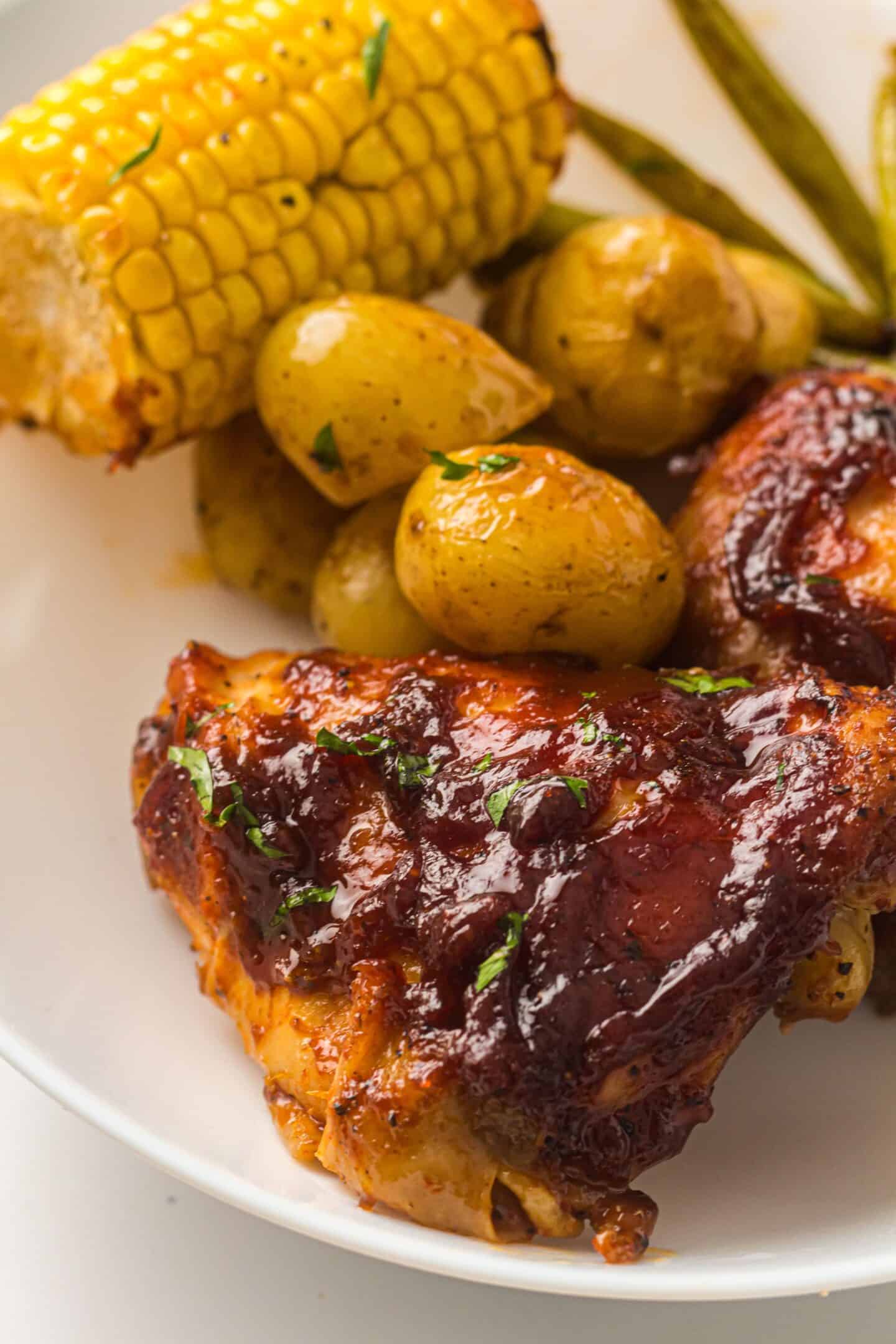 Plate with BBQ Chicken Thighs, roasted baby potatoes, green beans, and a piece of corn on the cob—all cooked to perfection on a sheet pan.