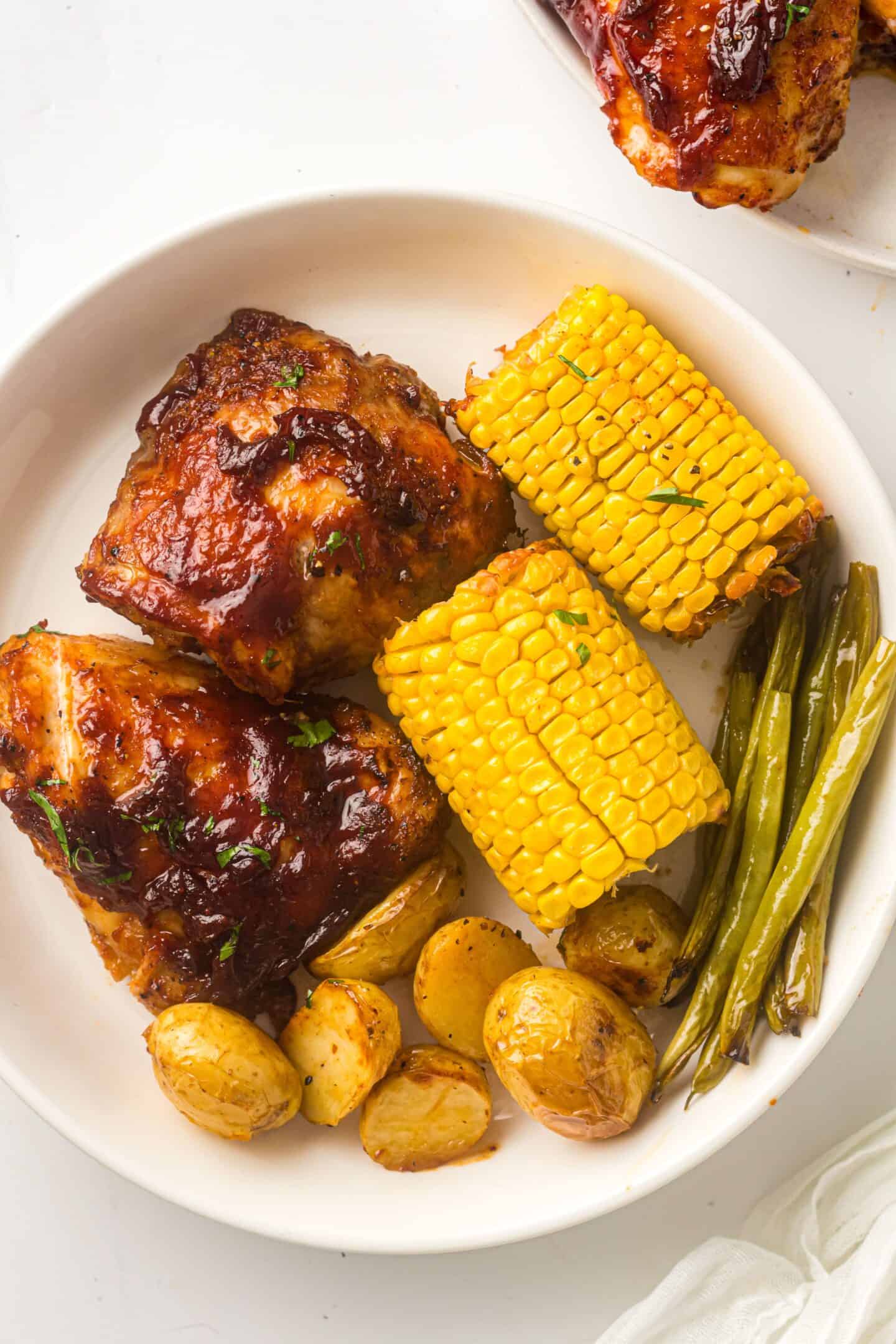 A plate featuring sheet pan BBQ chicken thighs, corn on the cob, roasted baby potatoes, and green beans.