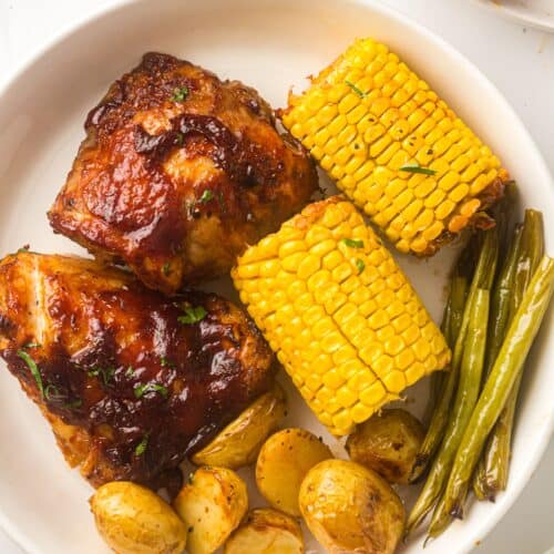 A plate featuring sheet pan BBQ chicken thighs, corn on the cob, roasted baby potatoes, and green beans.