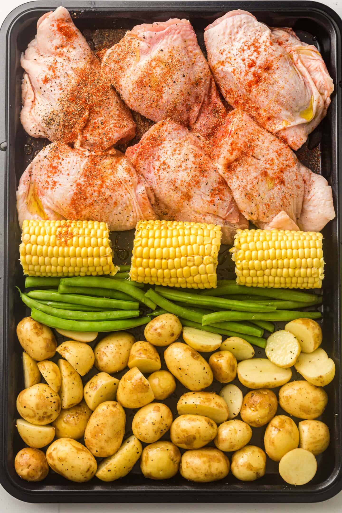 Sheet Pan BBQ Chicken Thighs are paired with fresh green beans, halved baby potatoes, and corn on the cob, all seasoned and arranged on a baking sheet, ready for roasting.
