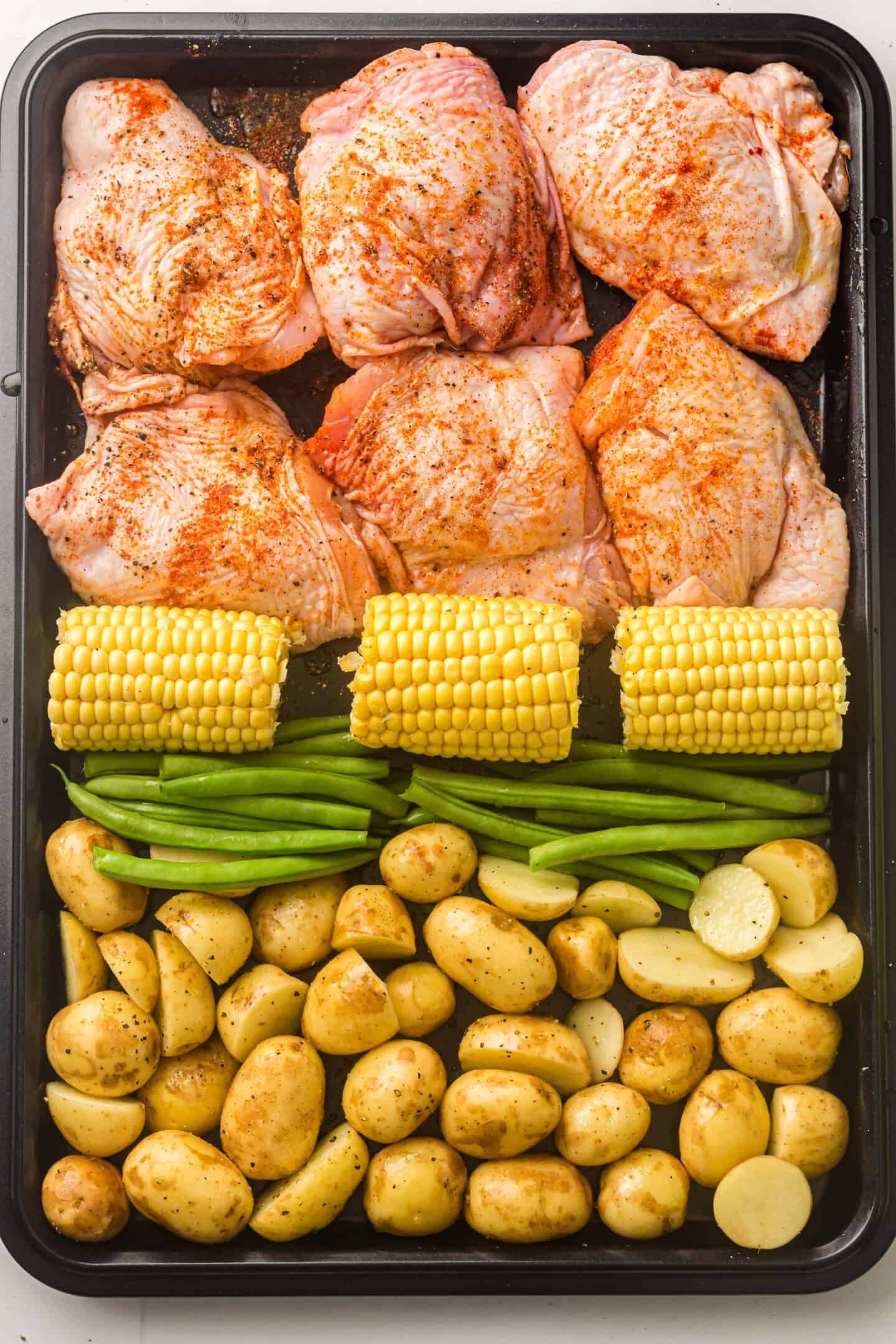 A sheet pan holds seasoned raw BBQ chicken thighs, corn on the cob, green beans, and halved baby potatoes, each arranged in separate sections for easy prep and roasting.