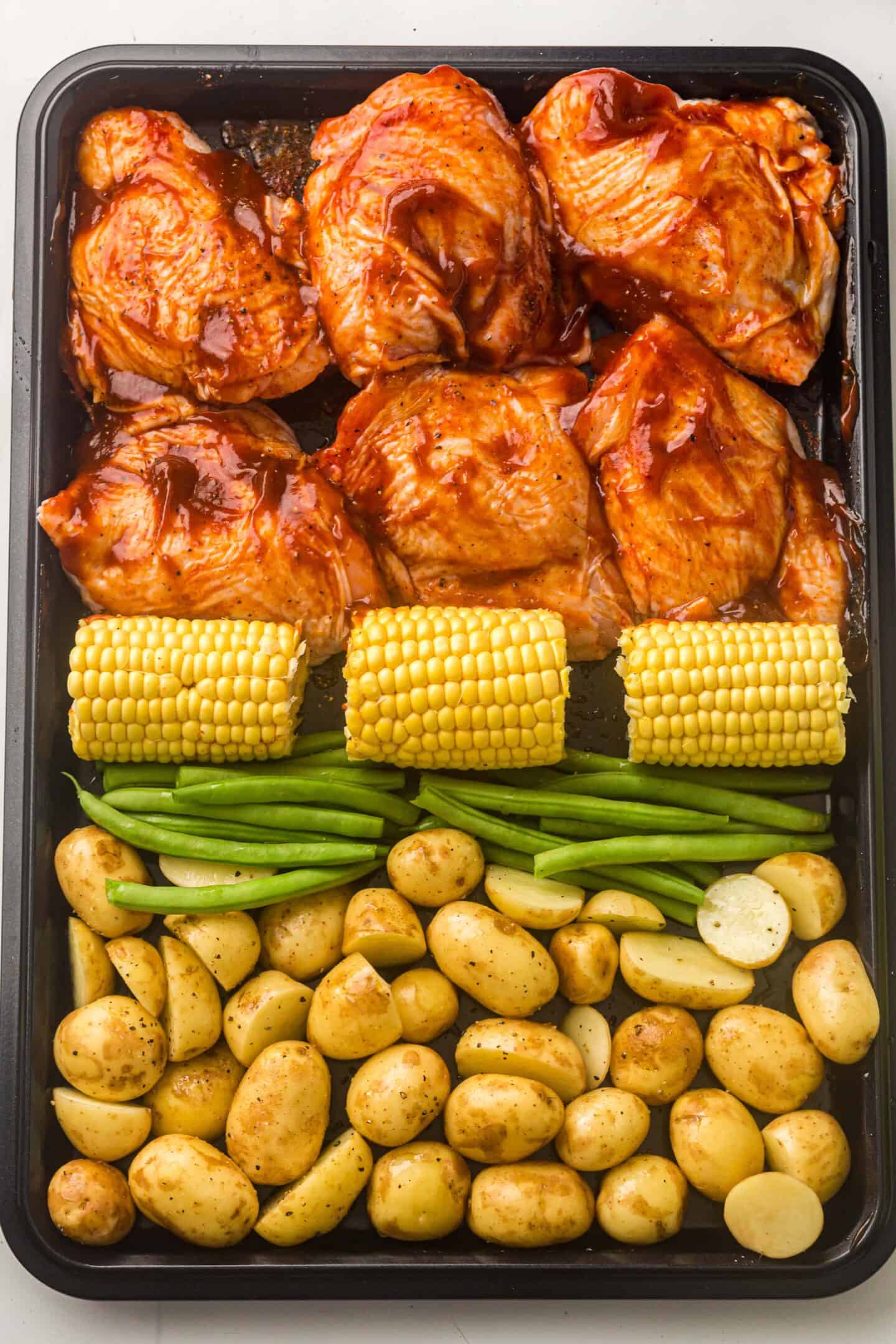 Sheet pan with raw BBQ chicken thighs, corn on the cob, green beans, and halved baby potatoes arranged in rows, ready for roasting—a delicious one-pan meal idea for your PLR content.