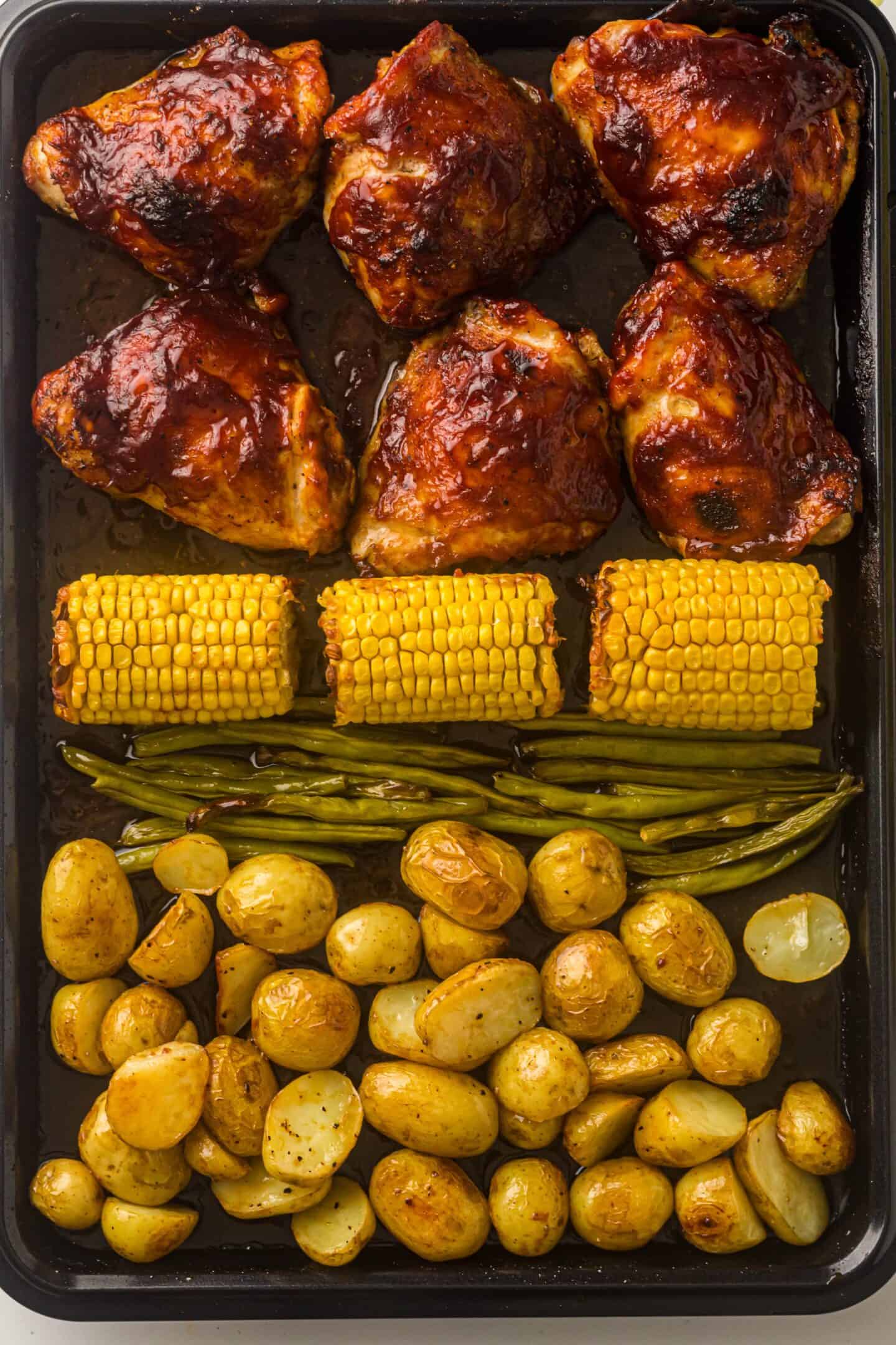 Sheet Pan BBQ Chicken Thighs baked with barbecue sauce, corn on the cob, green beans, and seasoned baby potatoes neatly arranged for a complete meal.