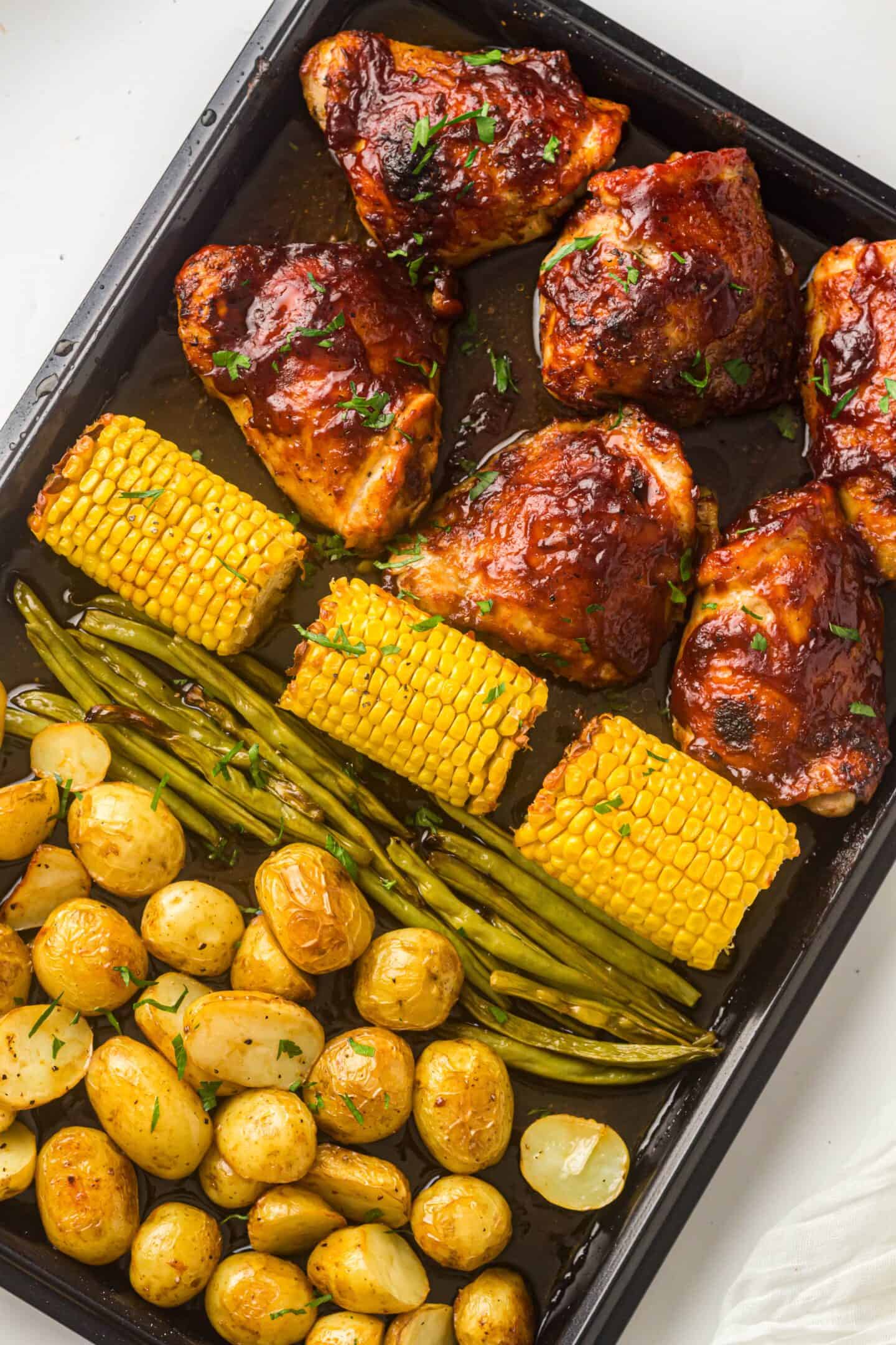 A sheet pan filled with roasted BBQ chicken thighs, corn on the cob, green beans, and baby potatoes, all garnished with fresh herbs.