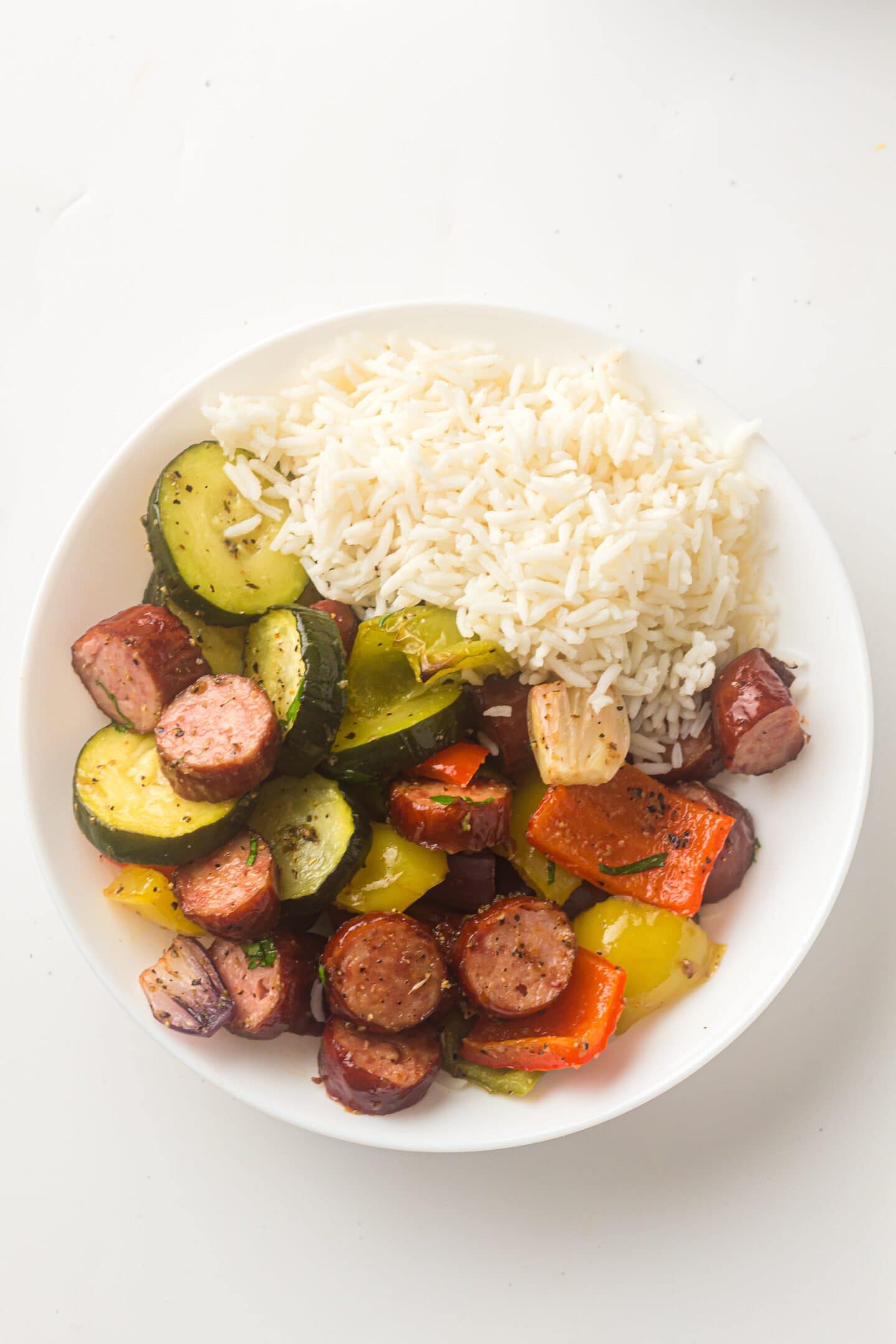 A white plate with white rice, sliced chicken sausage, and sheet pan roasted vegetables like zucchini, bell peppers, and garlic cloves.