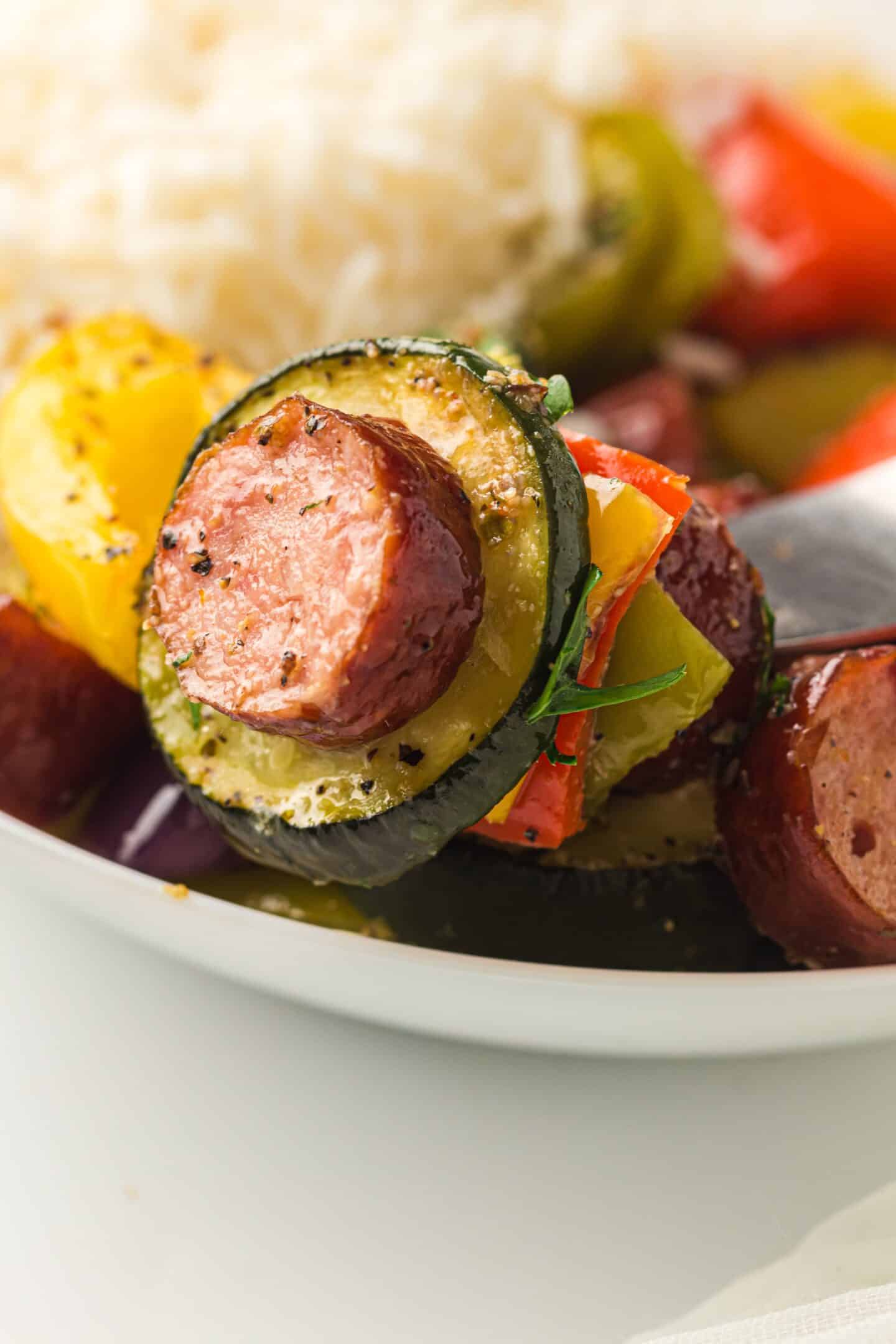 Sheet pan chicken sausage, zucchini, and bell peppers are served on a white plate with rice in the background, featuring perfectly roasted vegetables for a wholesome meal.