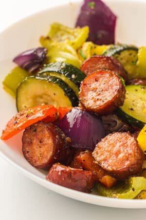 A white plate containing chicken sausage, zucchini, red onions, yellow and red bell peppers—all seasoned and roasted vegetables cooked on a sheet pan.