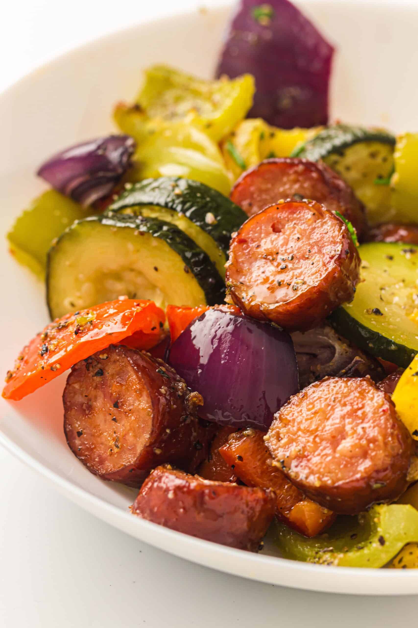 A white plate containing chicken sausage, zucchini, red onions, yellow and red bell peppers—all seasoned and roasted vegetables cooked on a sheet pan.