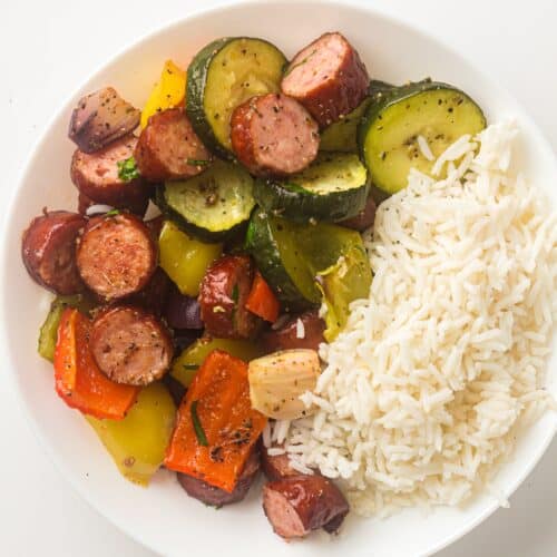 A plate with white rice, roasted vegetables, sliced chicken sausage, zucchini, red and yellow bell peppers, and onions on a white background.