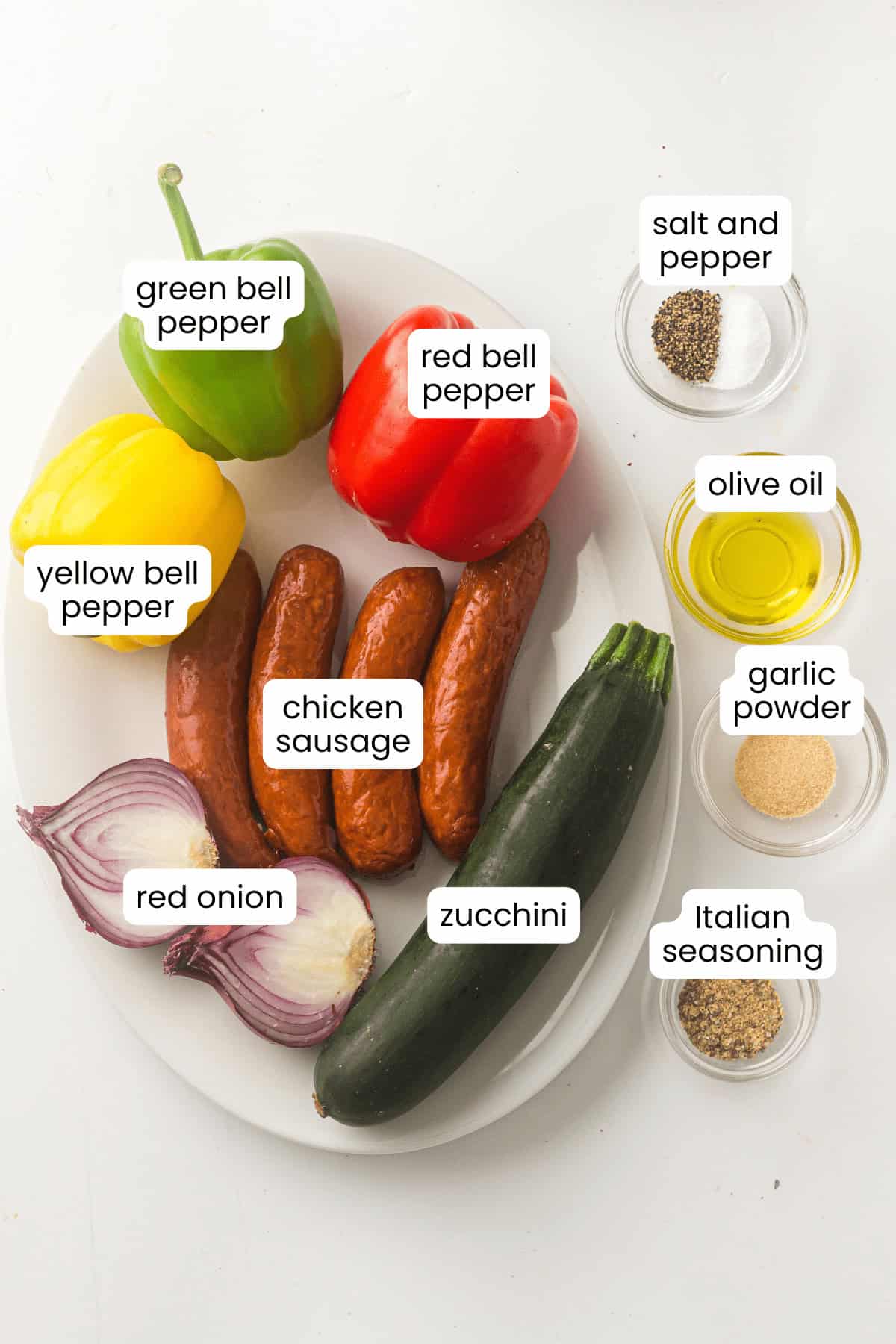 A white plate with sheet pan roasted vegetables—green, yellow, and red bell peppers, chicken sausage, red onion, and zucchini. Small bowls of salt and pepper, olive oil, garlic powder, and Italian seasoning sit beside the plate.