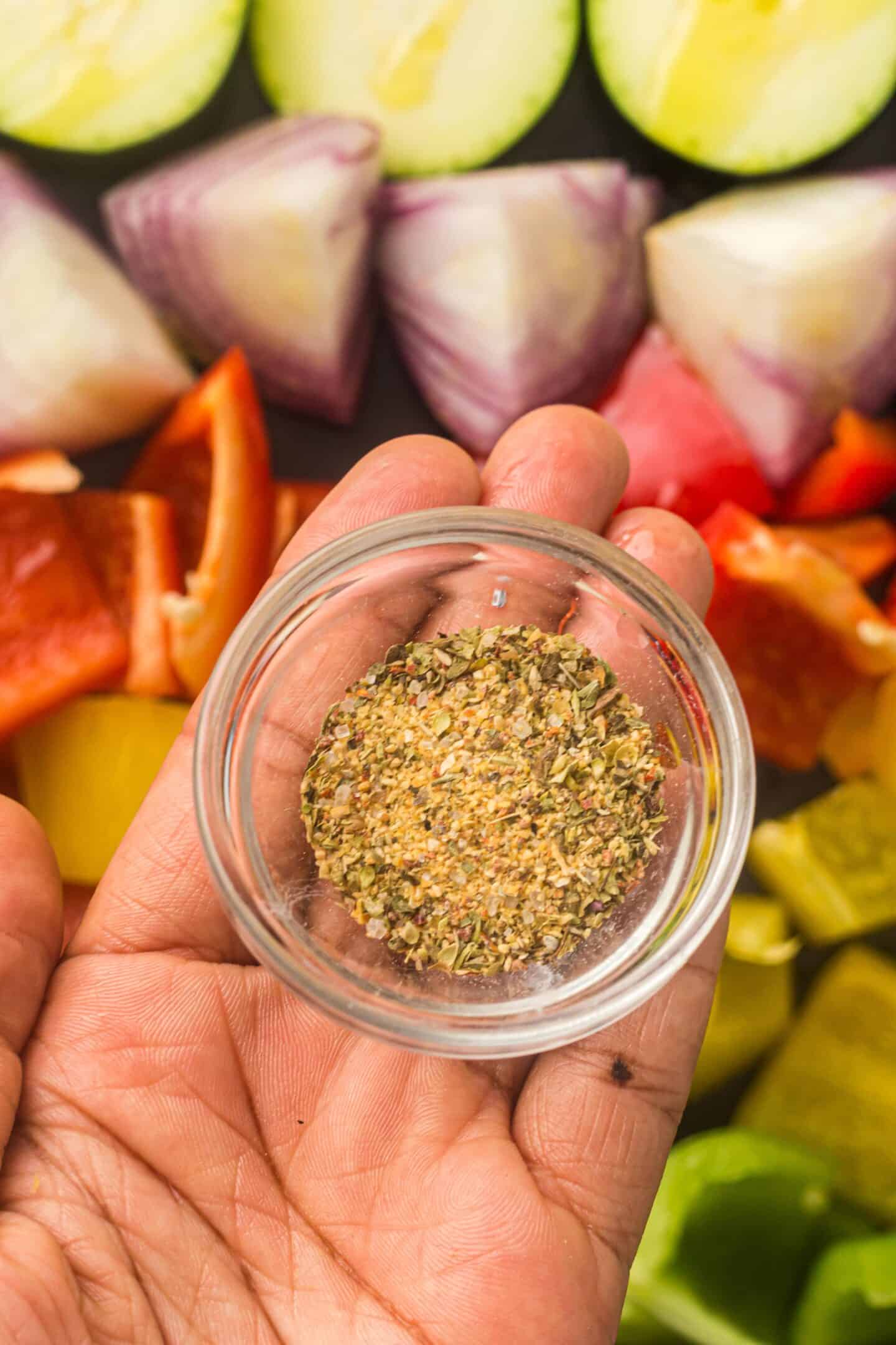 A hand holds a small glass bowl of mixed dried spices above a background of sheet pan roasted vegetables, including onions, peppers, zucchini, and slices of chicken sausage.