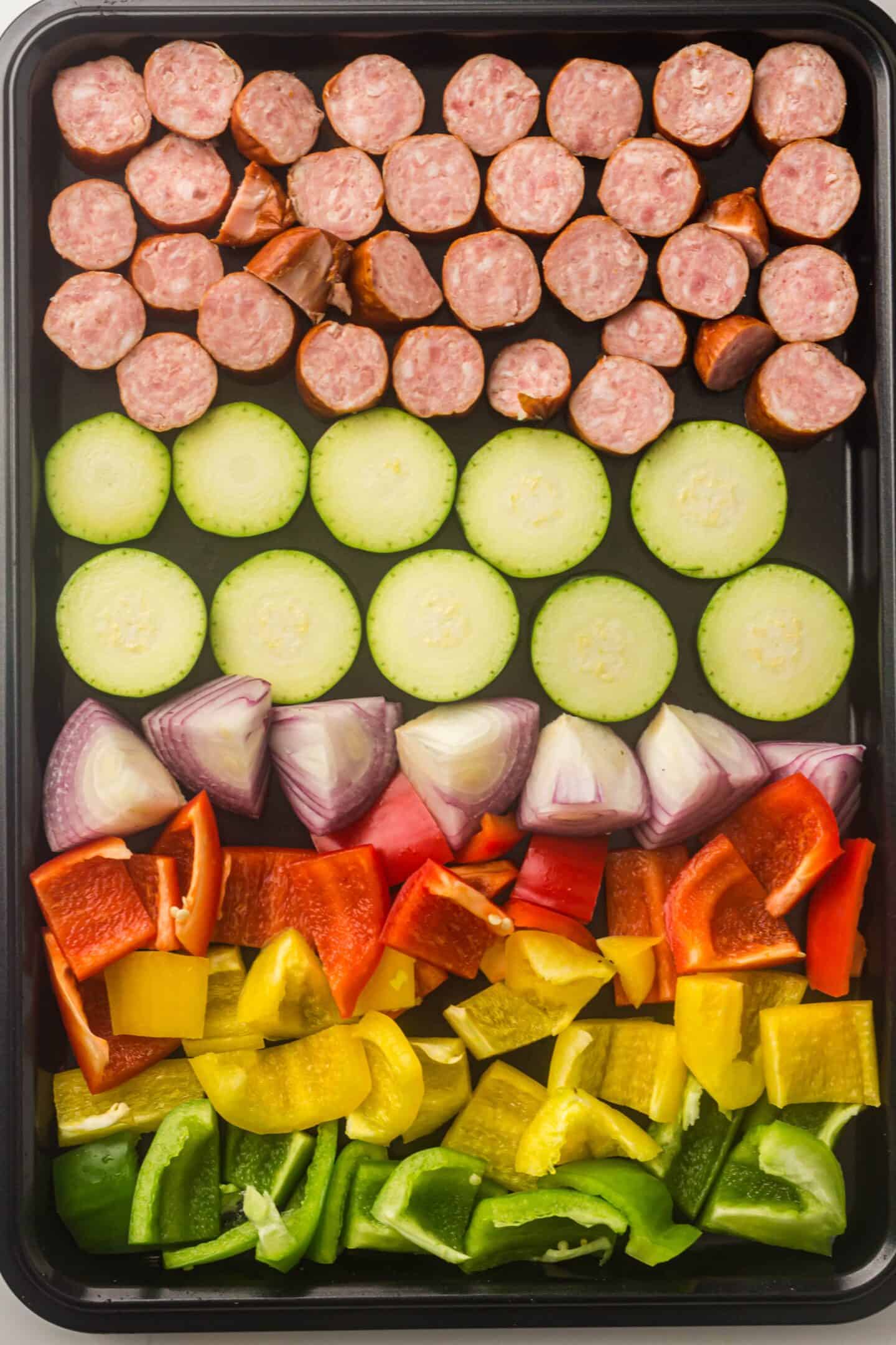 A sheet pan with rows of sliced chicken sausage, zucchini, red onion, and chopped red, yellow, and green bell peppers arranged neatly for roasted vegetables.