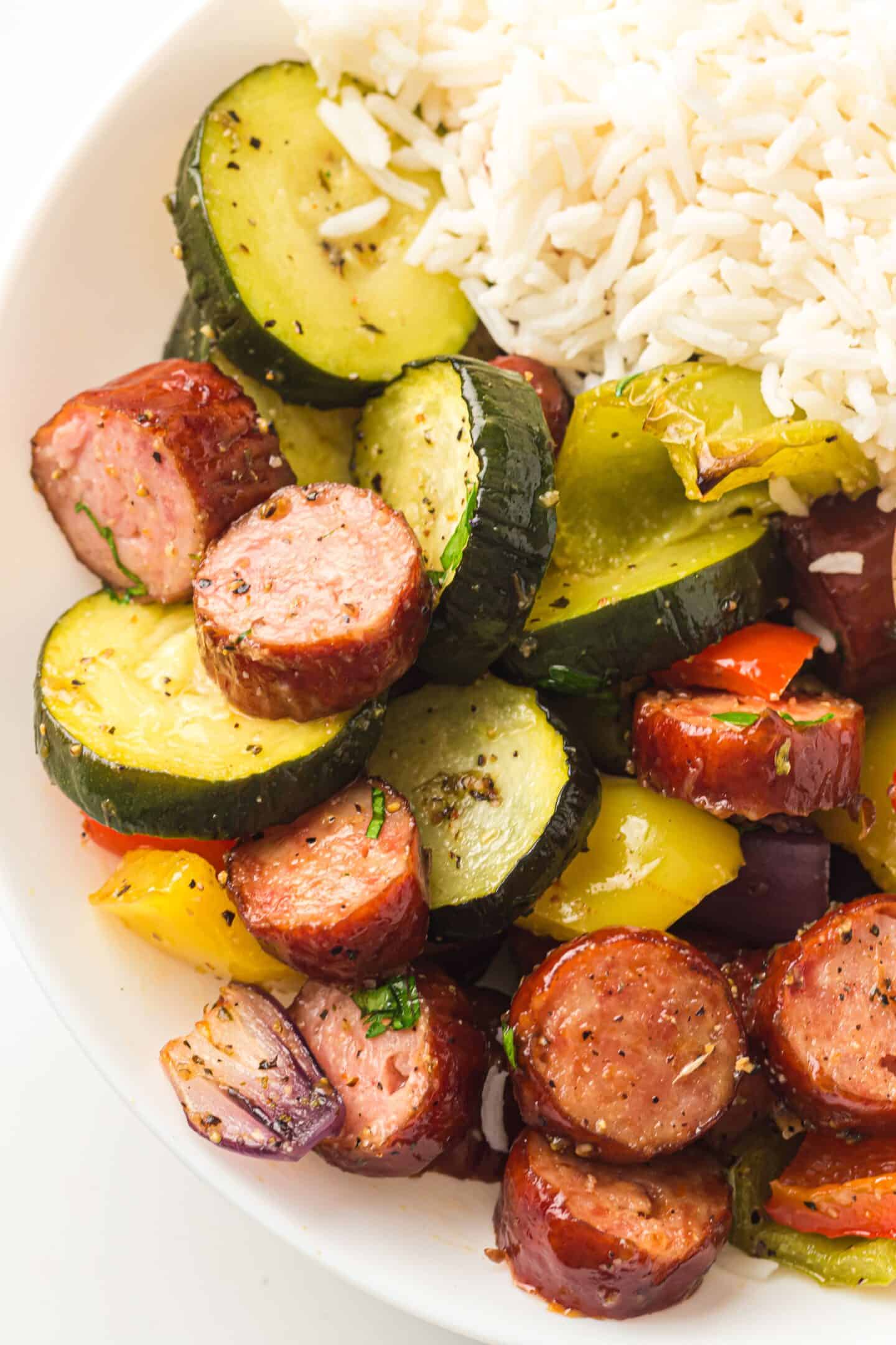 Sliced sheet pan chicken sausage, roasted vegetables like zucchini, bell peppers, and onions, all served with white rice on a white plate.