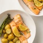Two white plates each with sheet pan salmon fillets topped with lemon slices, accompanied by roasted potatoes and fresh asparagus spears.