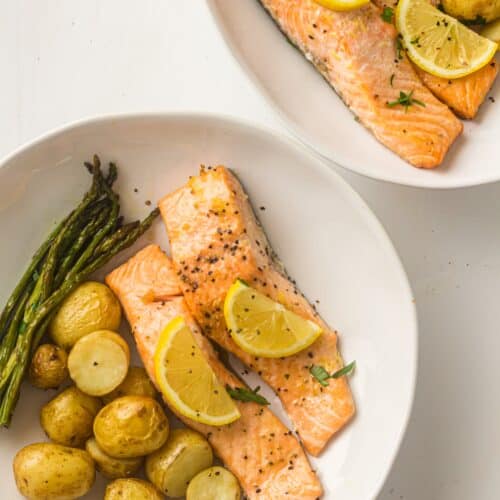 Two white plates each with sheet pan salmon fillets topped with lemon slices, accompanied by roasted potatoes and fresh asparagus spears.